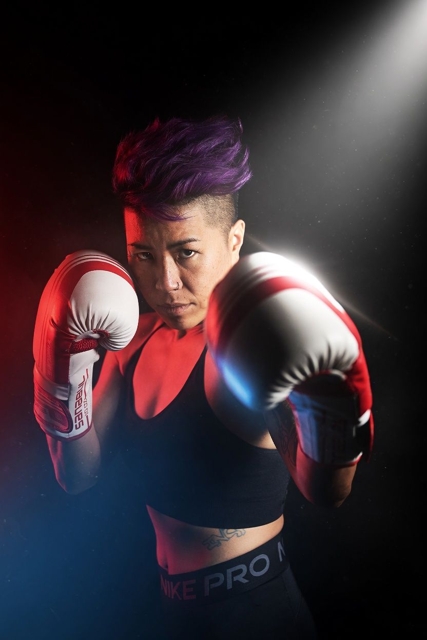 Female boxer with purple hair in Nike Pro gear poses aggressively in red and white boxing gloves under dramatic spotlight.