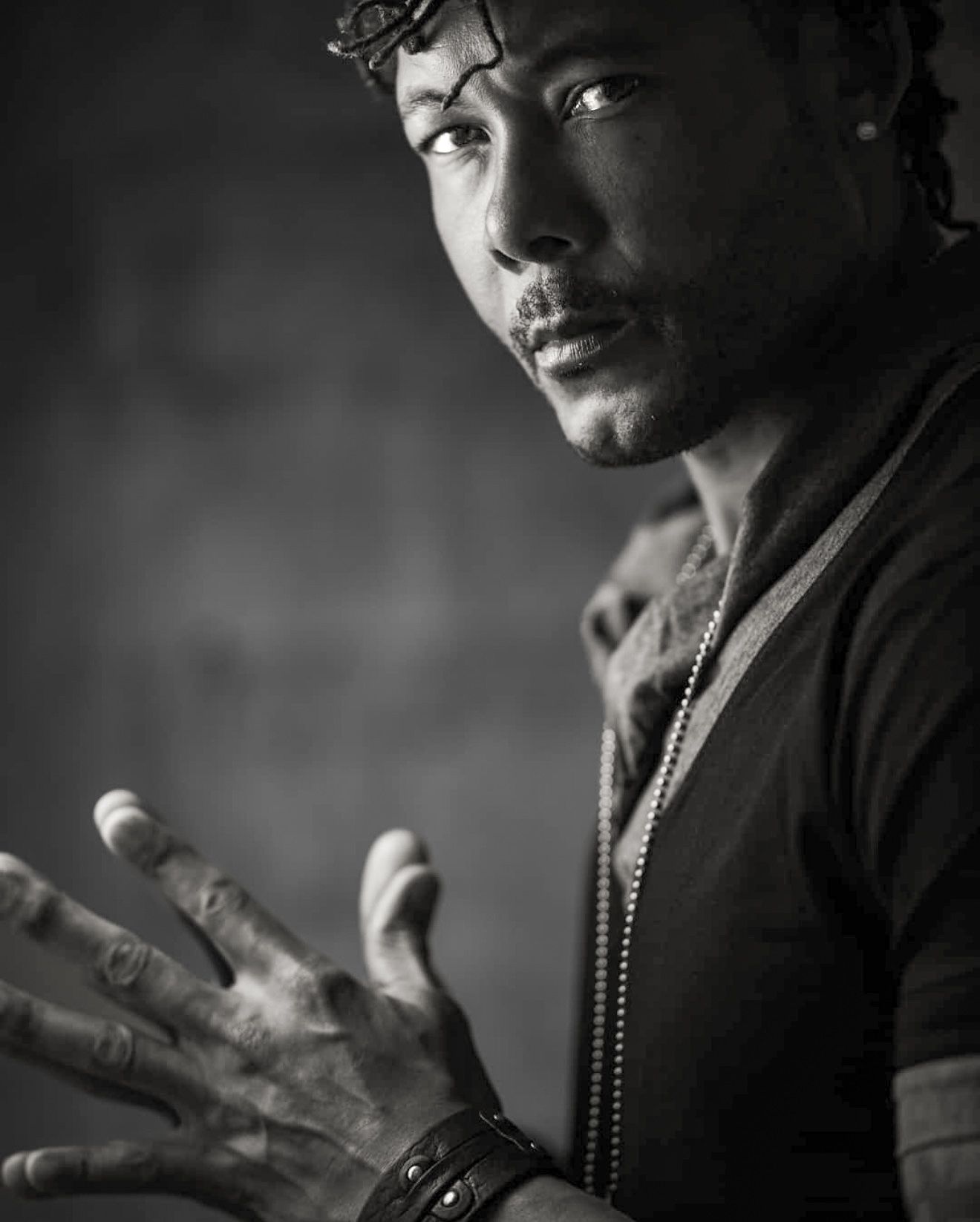 Black and white artistic portrait of man with raised hand wearing bracelet and necklace.