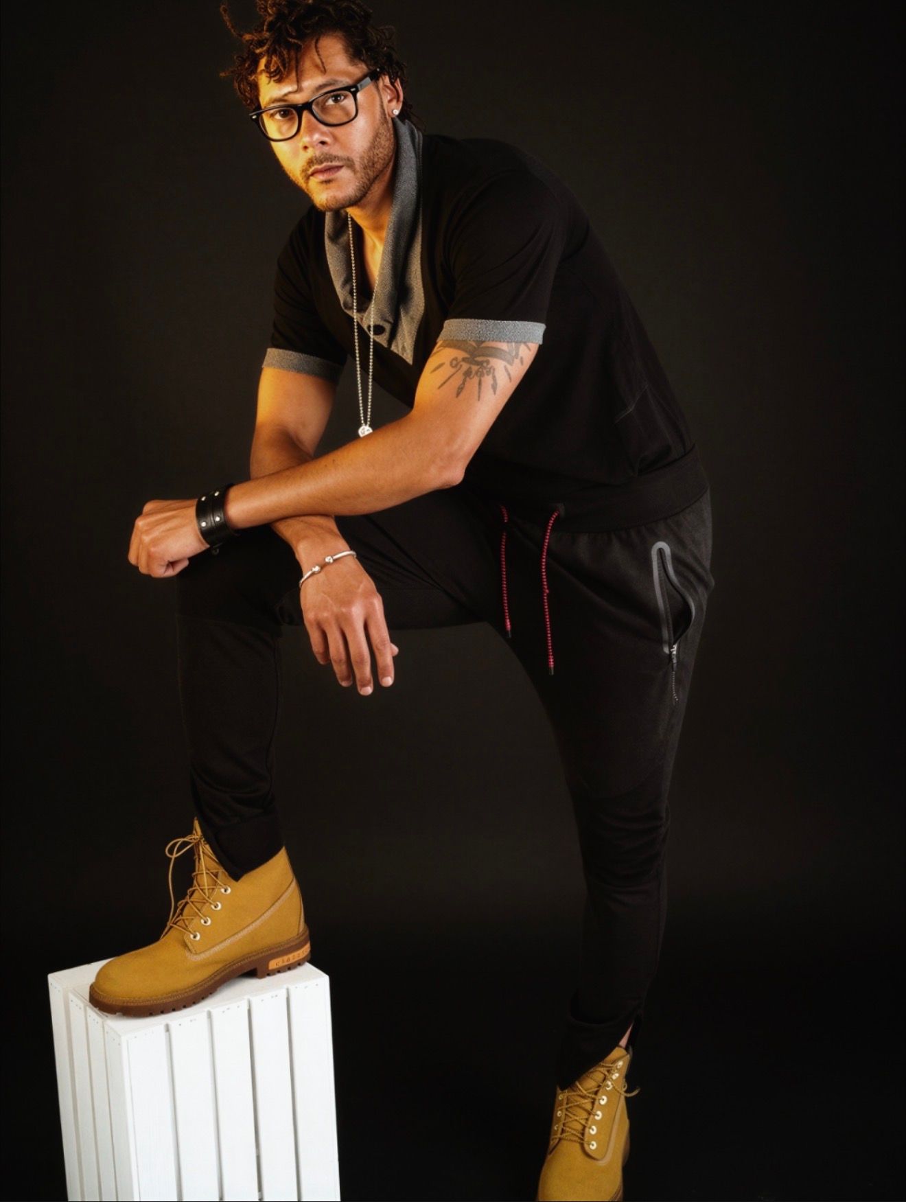 Stylish man in black outfit with tan Timberland boots and glasses posing confidently against dark background.