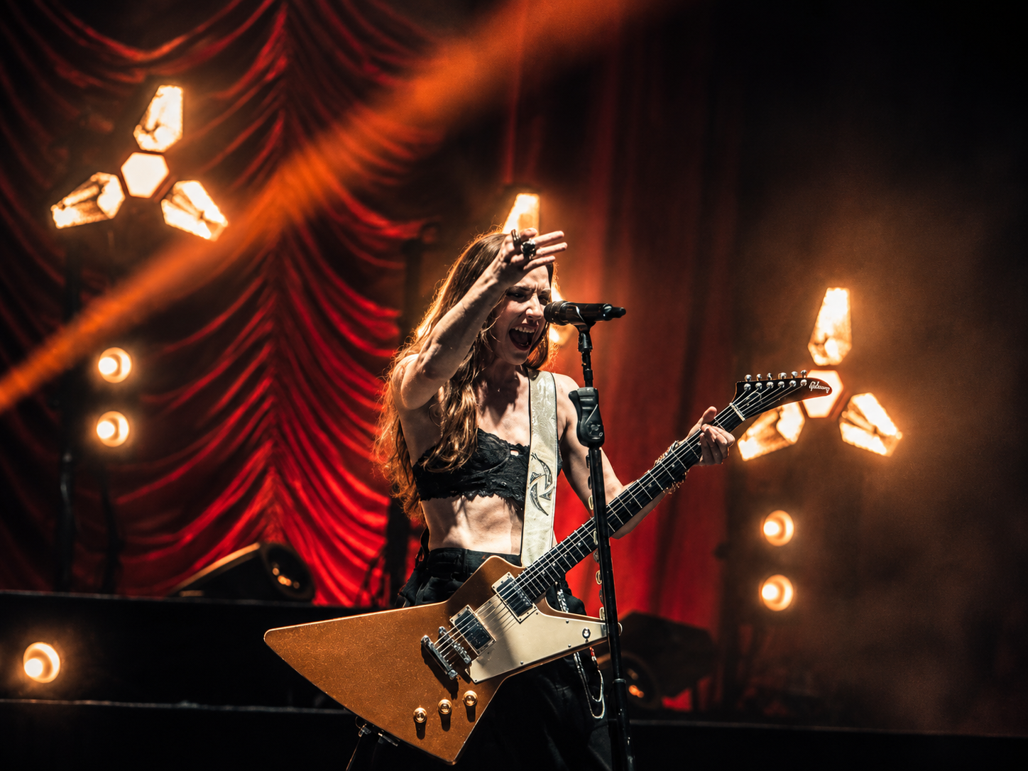 Female rock guitarist performs on stage with Gibson Explorer guitar, red curtain backdrop, and dramatic stage lighting.