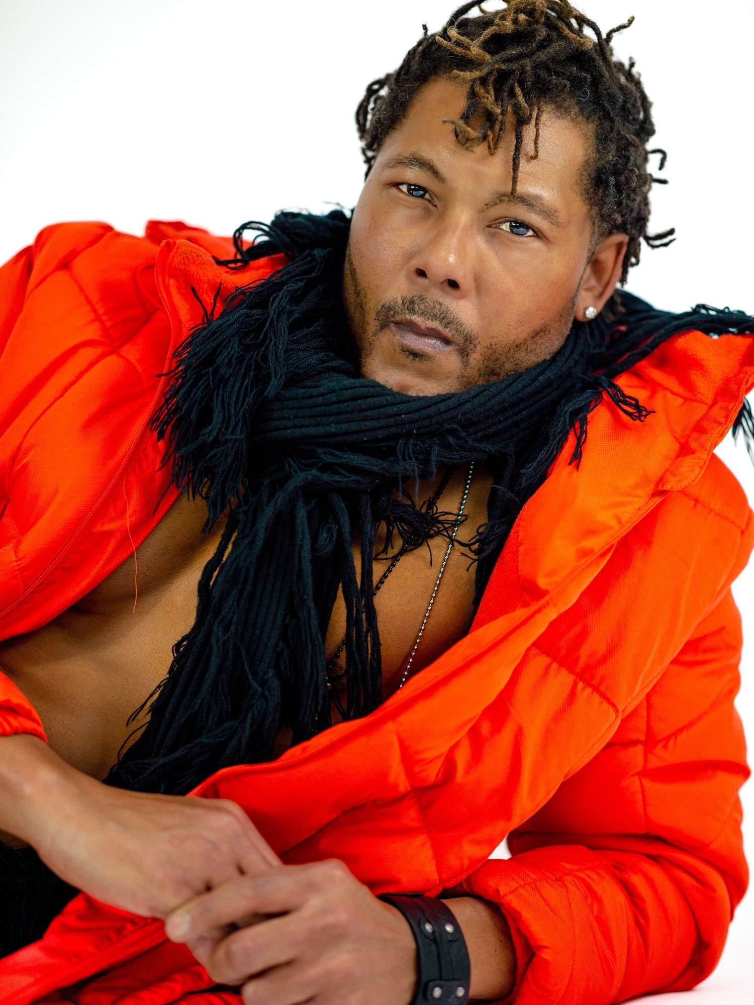 Man in orange puffer jacket and black fringe scarf posing confidently against white background.