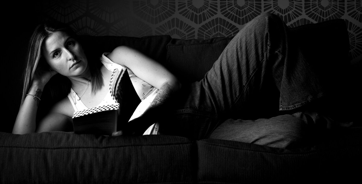 Woman reading a book while lying on a dark sofa, captured in moody black and white photography.