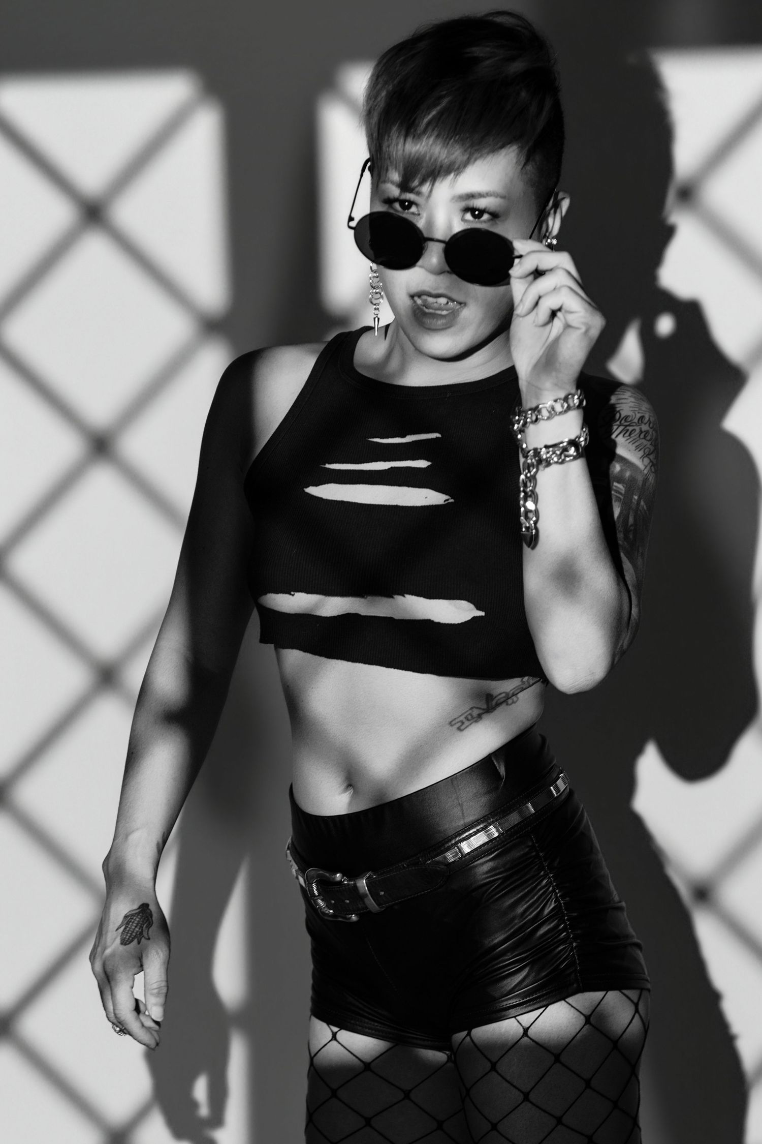 Black and white photo of a woman in edgy outfit with fishnet stockings, crop top, and sunglasses posing confidently.