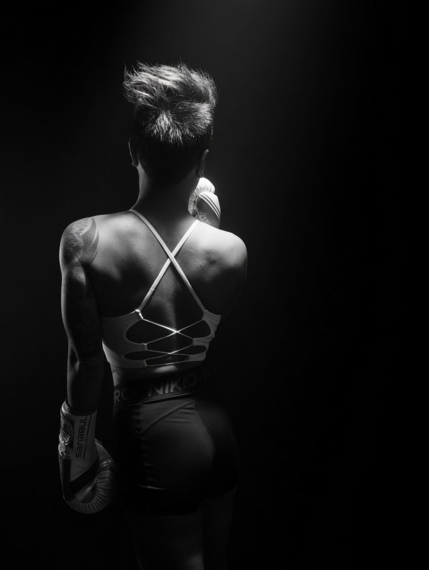 Black and white photo of a muscular woman boxer seen from behind, wearing a sports bra and boxing gloves.