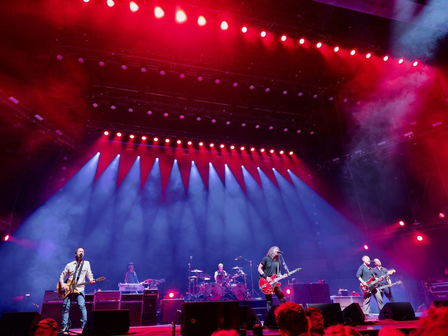 Rock band performs on a dramatic stage with red and blue spotlights and smoke effects at a large concert venue.
