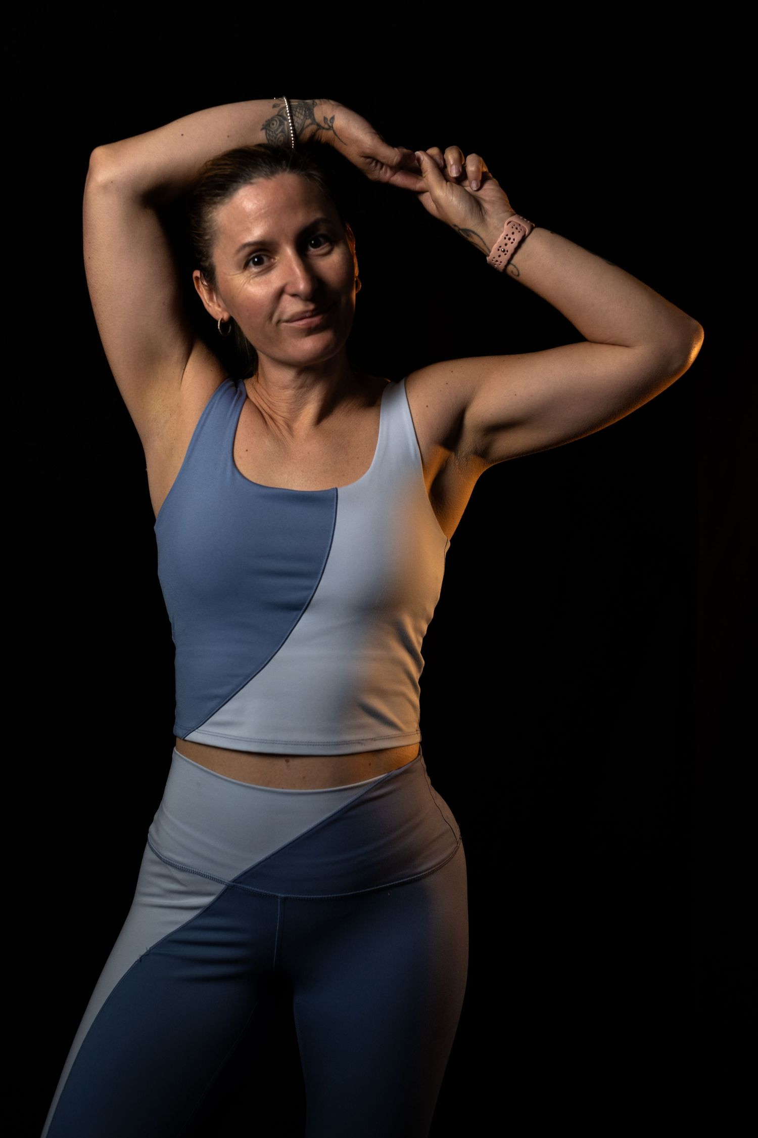 Fit woman in blue and gray athletic wear poses confidently against dark background, arms raised, showing tattoos.