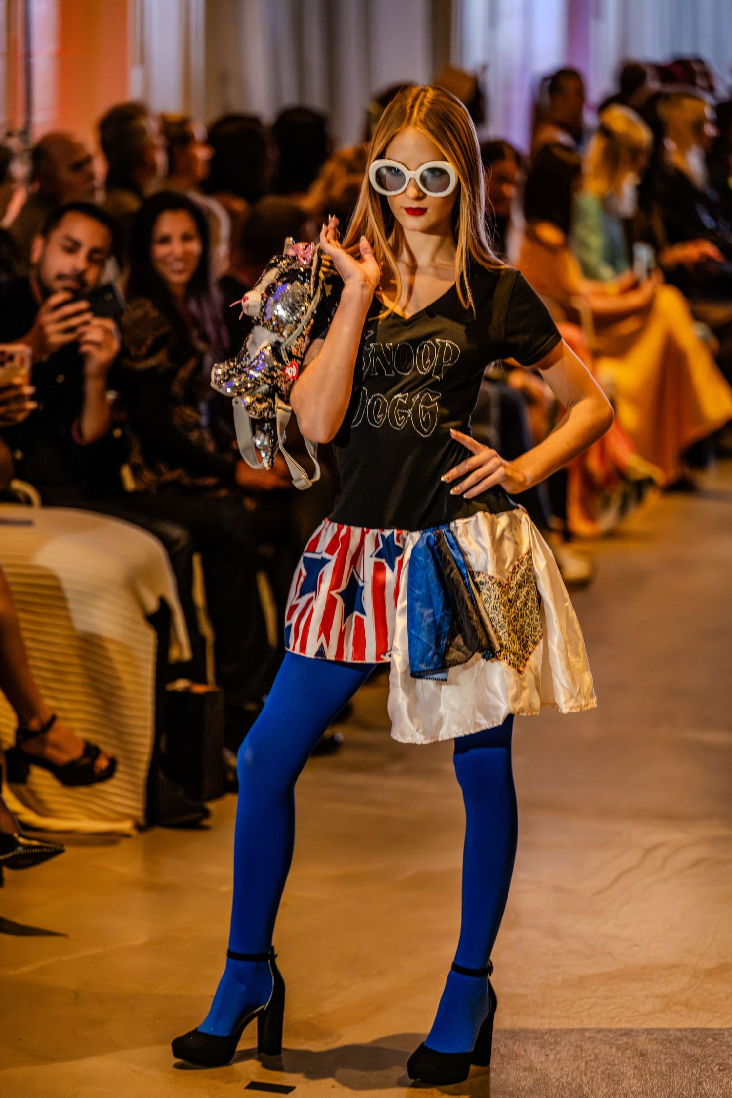Model wearing blue tights, a colorful patchwork skirt and white sunglasses on the Charlotte Fashion Week runway.