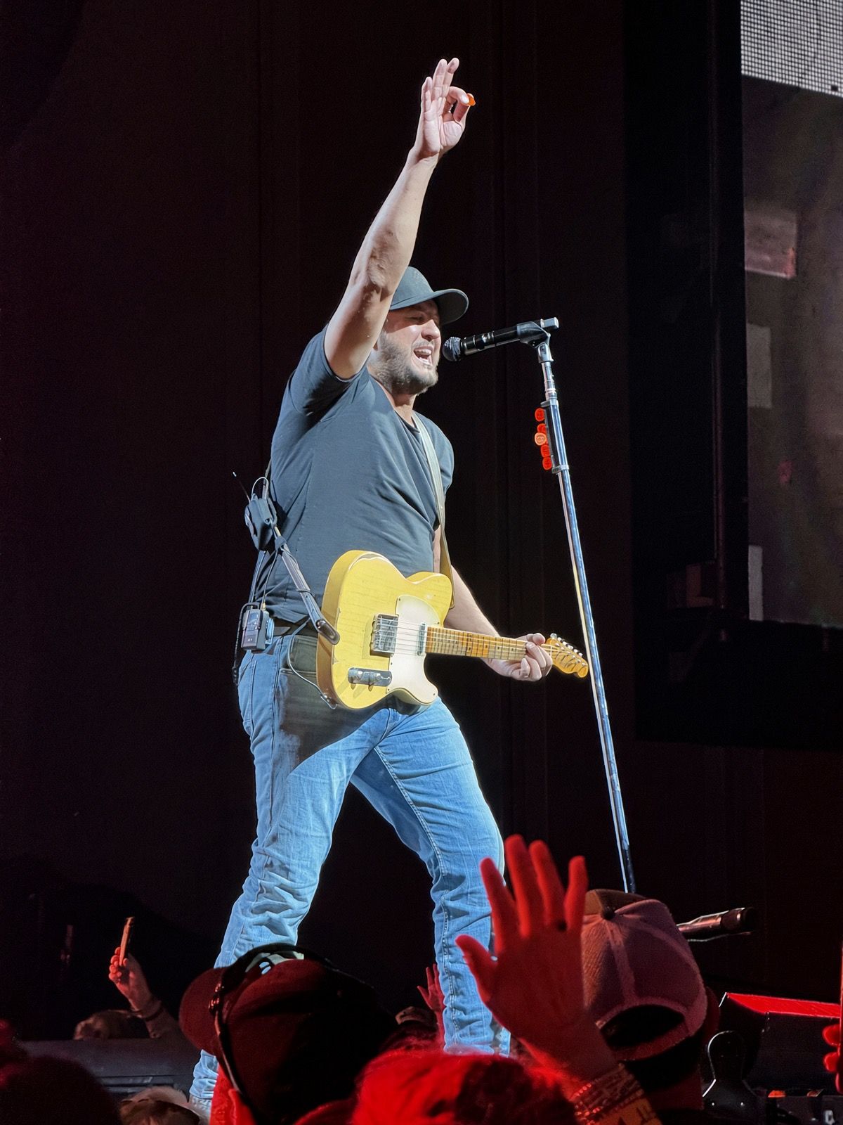 Male guitarist in jeans and cap raises hand while performing on stage with yellow electric guitar at live concert.