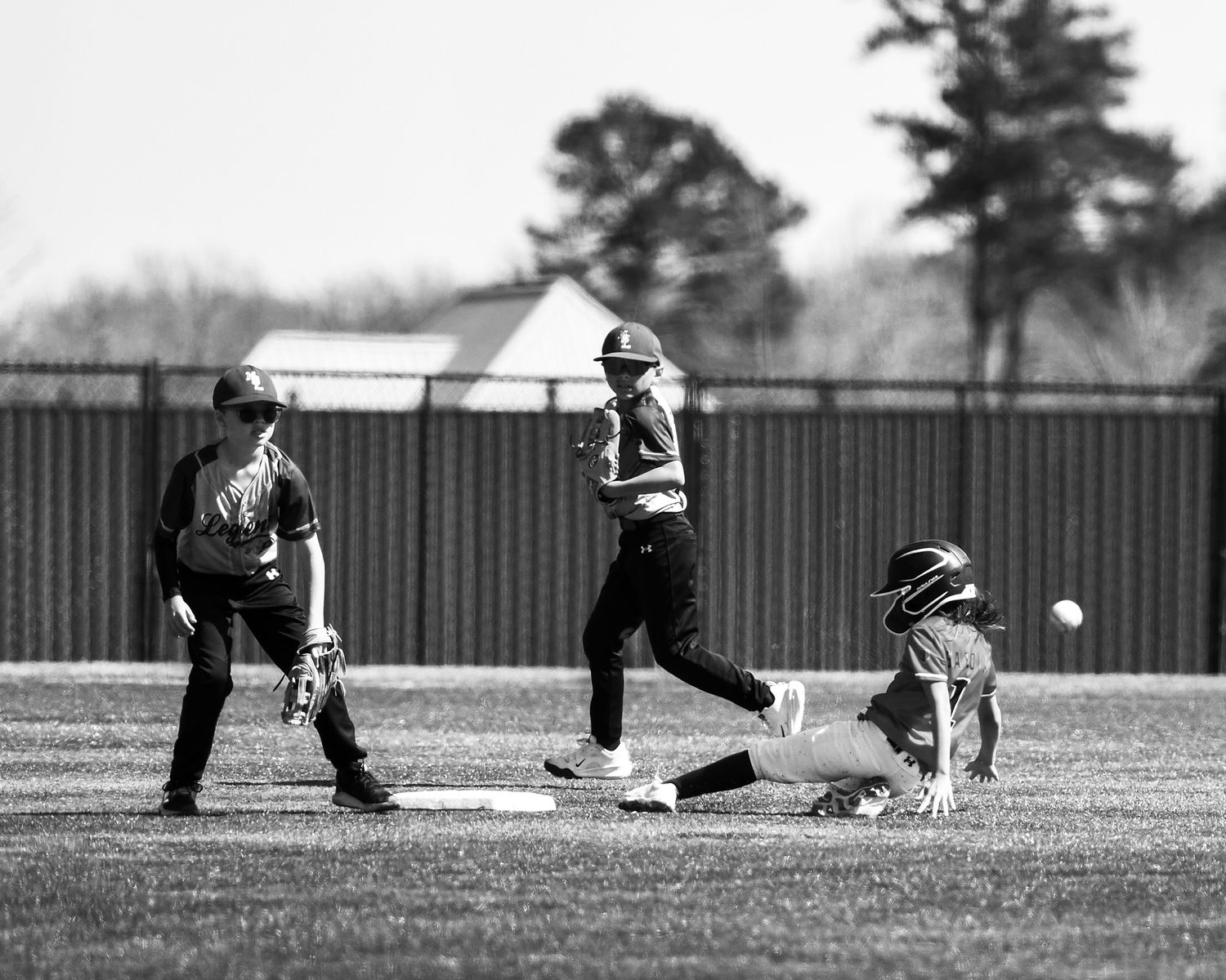 Black and white youth baseball game with a player sliding into second base as fielders await the throw.