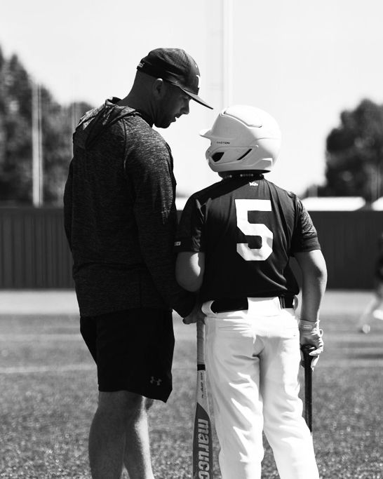 Black and white photo of a baseball coach instructing a young player wearing number 5 jersey and a batting helmet.