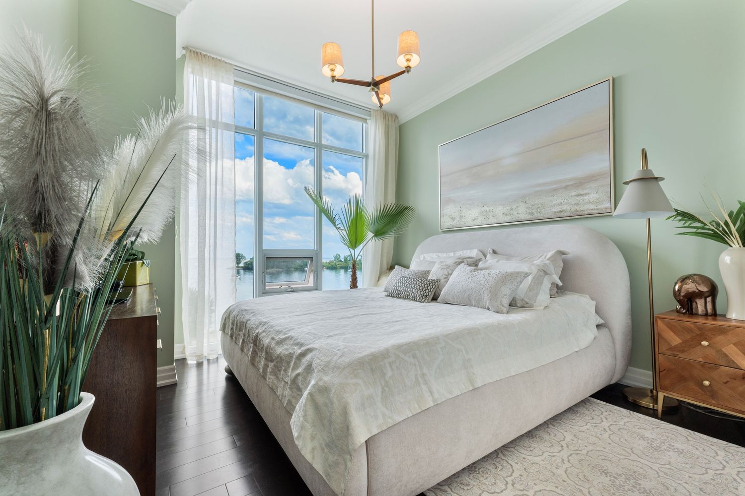 Elegant bedroom with sage green walls, upholstered bed, dark hardwood floors, chandelier, and floor-to-ceiling windows with water views.