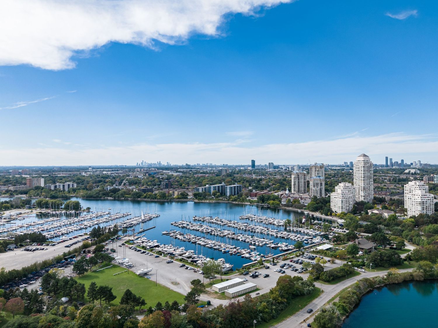 Aerial view of a marina with sailboats, waterfront park, and city skyline under a bright blue sky.