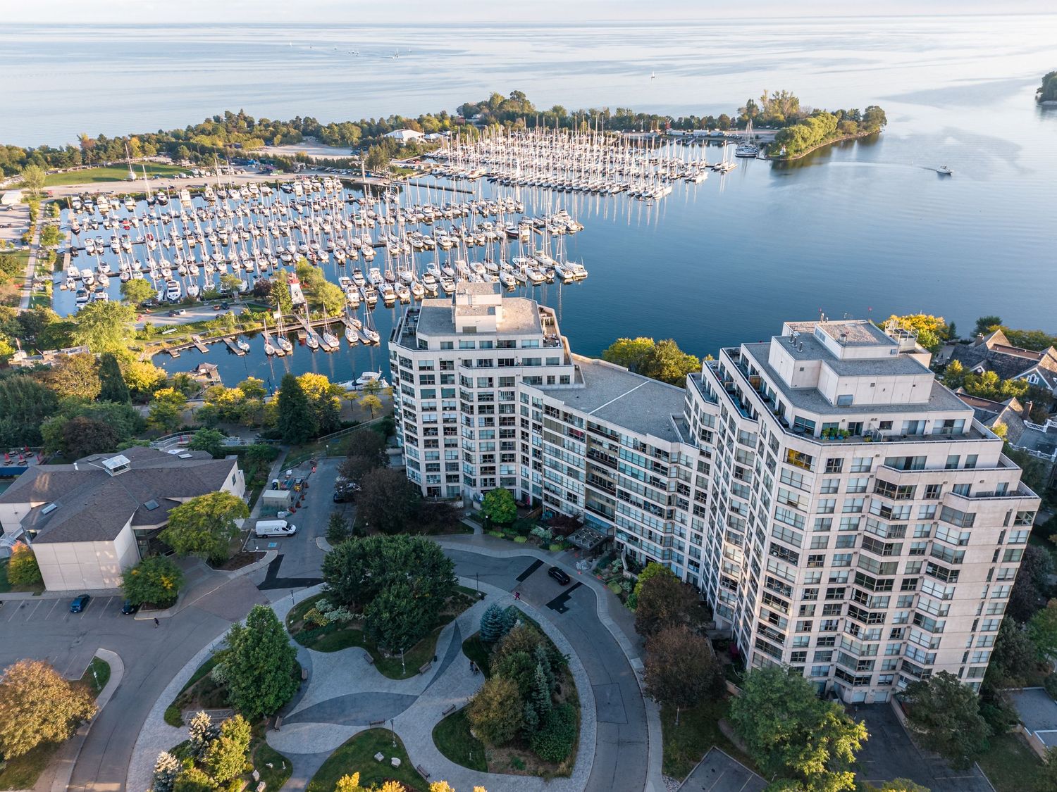 Aerial view of luxury waterfront condos beside a bustling marina with sailboats on calm blue water at sunset.