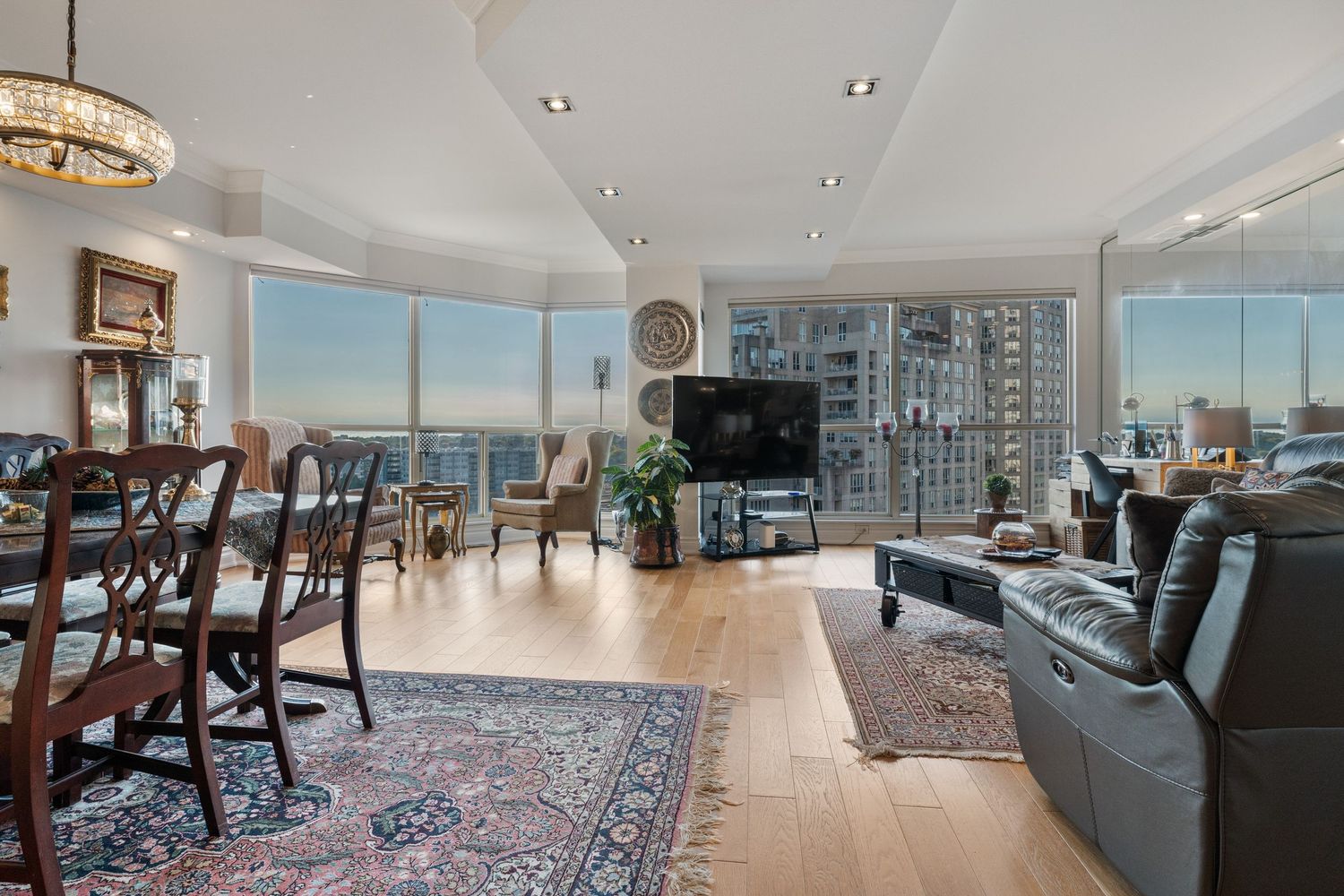 Spacious high-rise living room with hardwood floors, leather sofas, dining area, and panoramic city views through floor-to-ceiling windows.