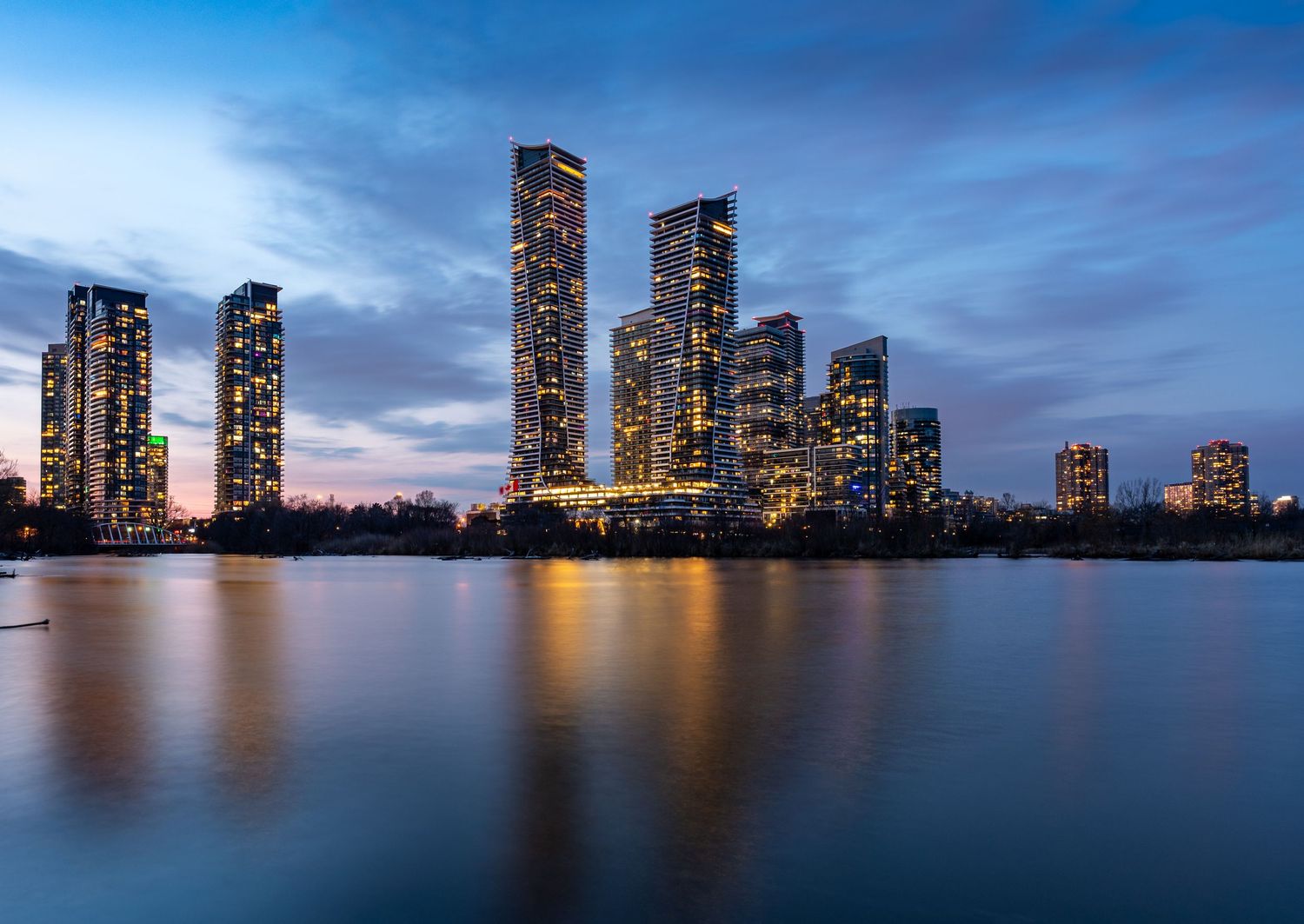 Twilight cityscape featuring tall illuminated skyscrapers reflecting golden light on a calm waterfront at Humber Bay Shores.