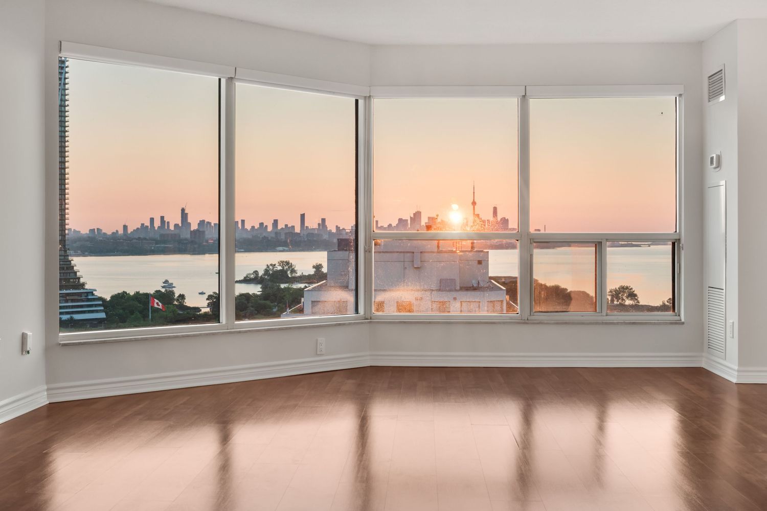 Empty room with hardwood floors and large bay windows showcasing a stunning city skyline and waterfront sunset view.
