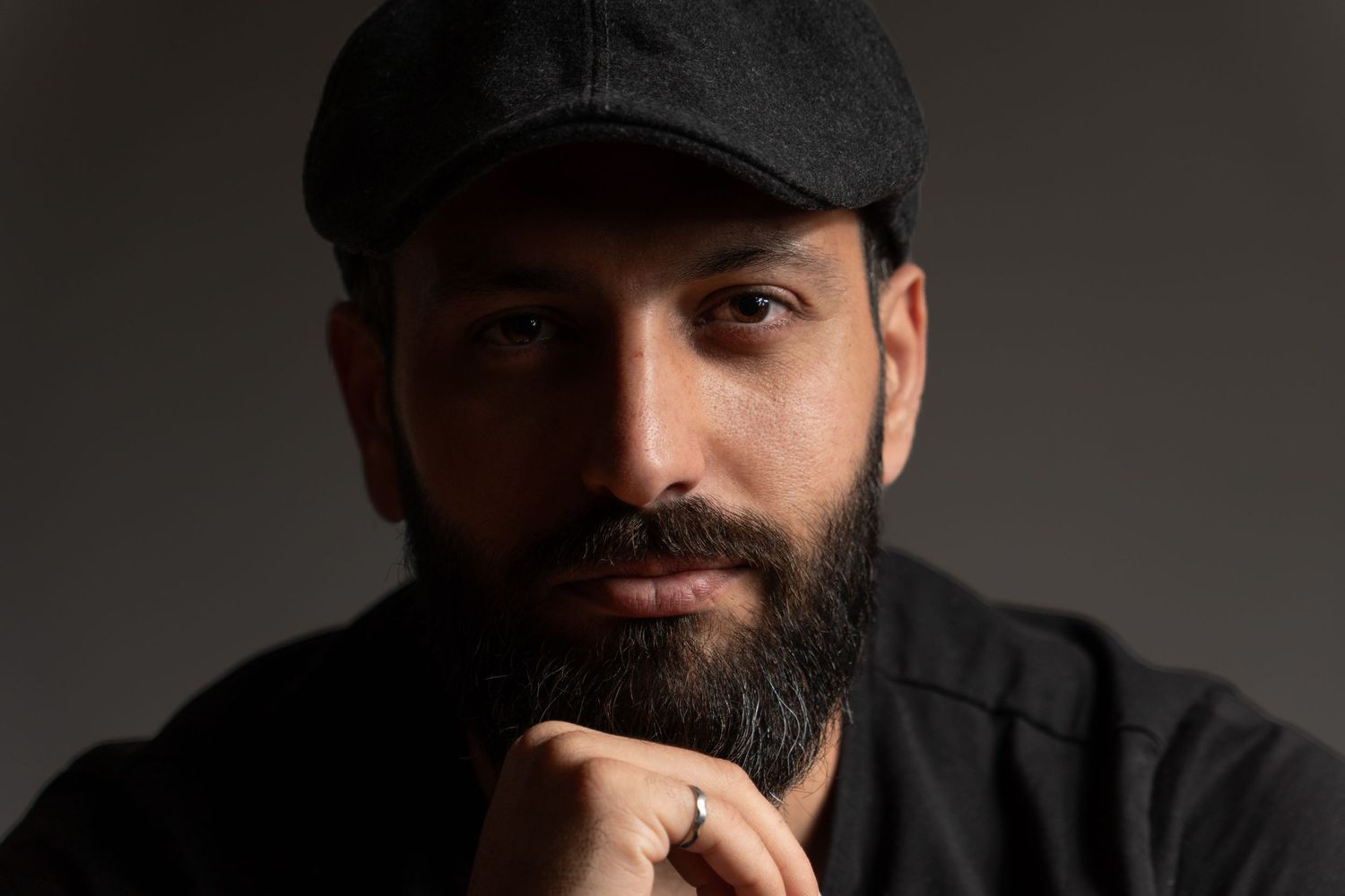 Portrait of a bearded man wearing a dark cap with hands clasped under chin against gray background.