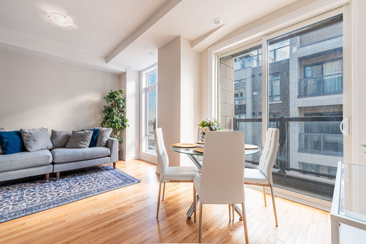 Bright dining area with white chairs, glass table, hardwood floors, and large windows overlooking city.