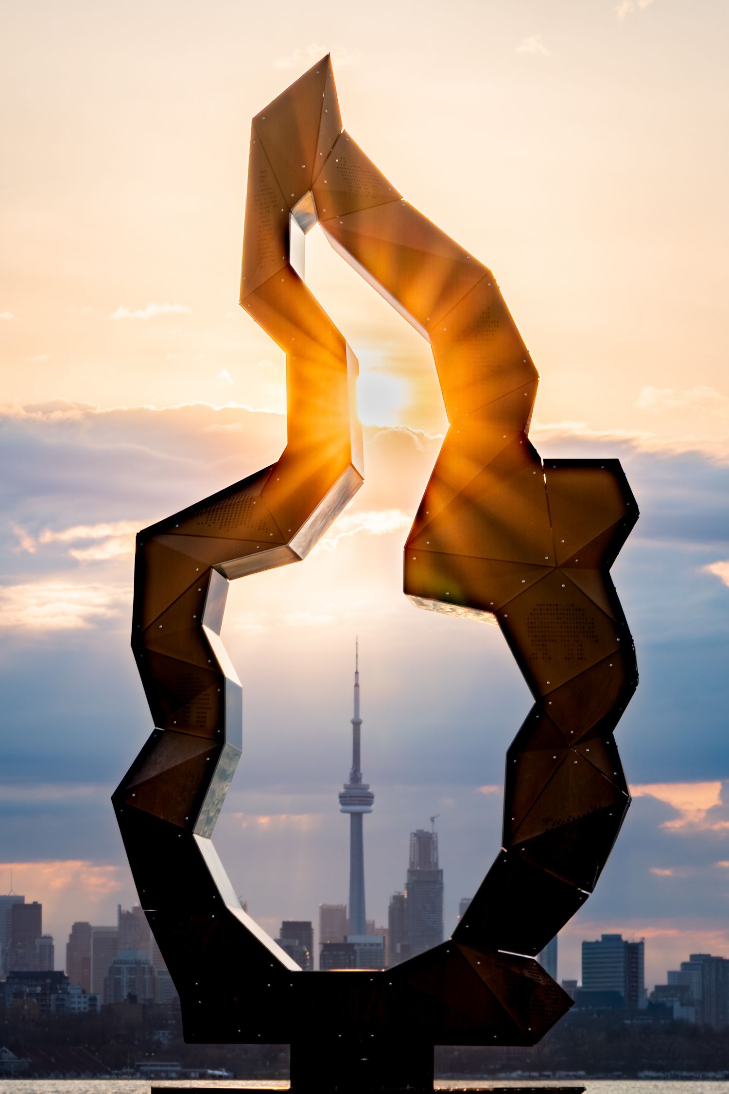 Abstract metal sculpture framing the CN Tower and Toronto skyline at golden sunset over the lake from Humber Bay Shores.
