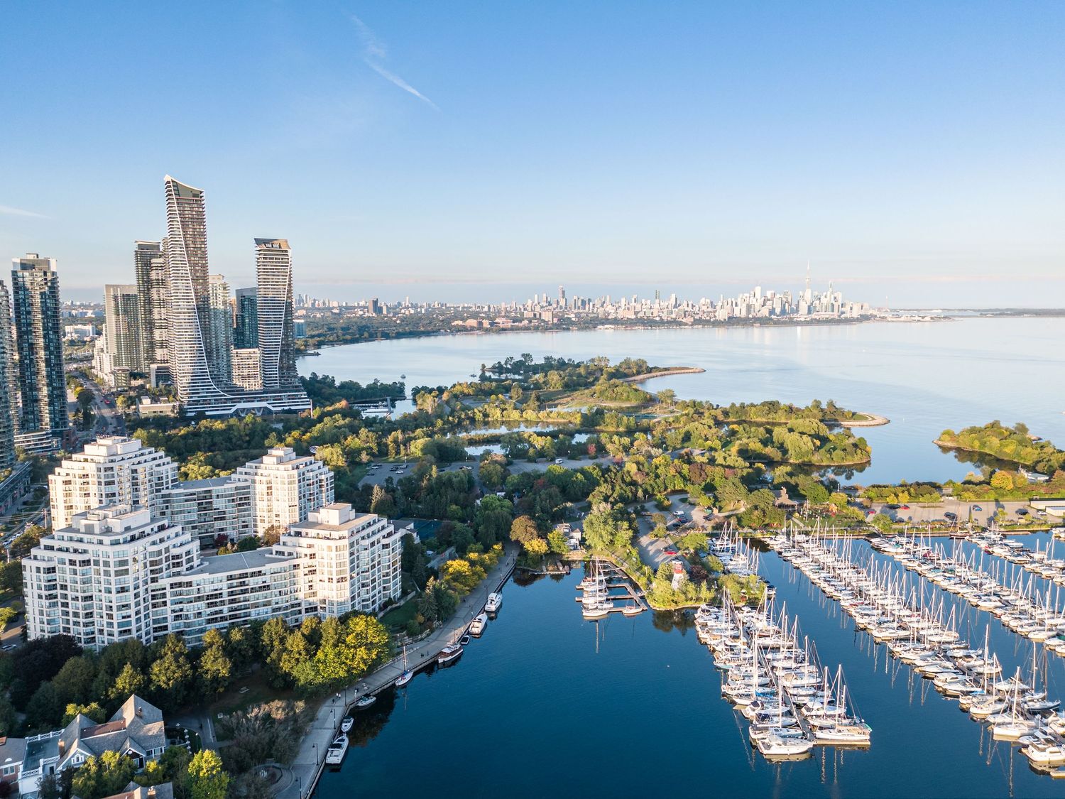 Aerial view of a marina with sailboats, lush green park, and modern skyscrapers along a calm waterfront on a sunny day.