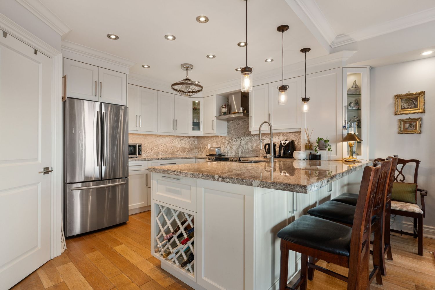 Elegant white kitchen with granite island, pendant lights, stainless steel fridge, wine rack, and hardwood floors.