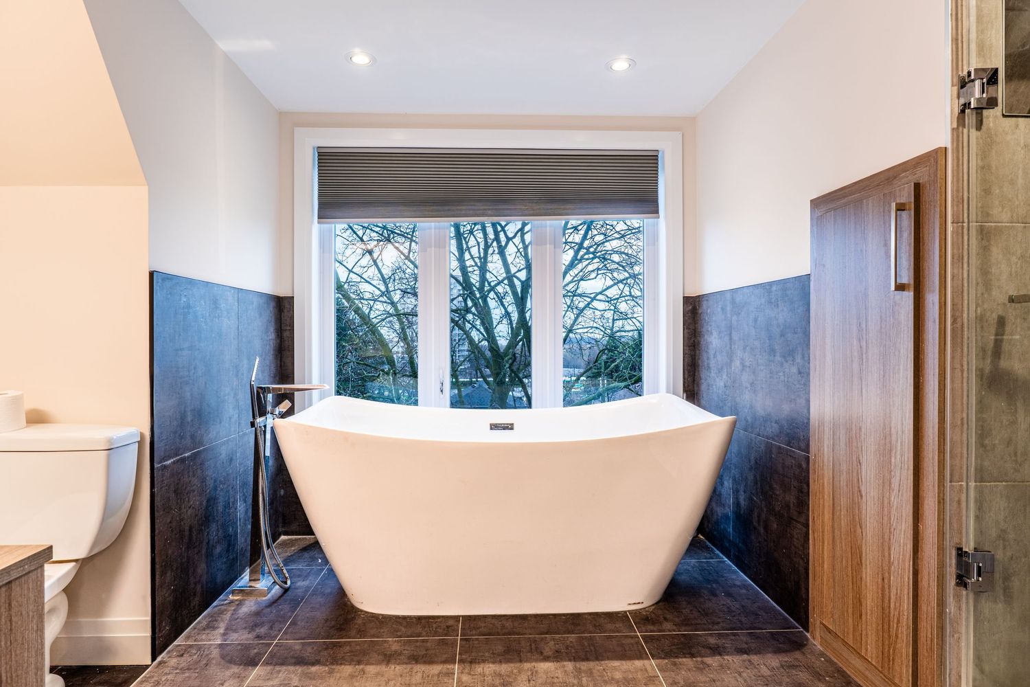 Luxury freestanding white soaking tub centered beneath a large window with blue tile walls and wood accents.