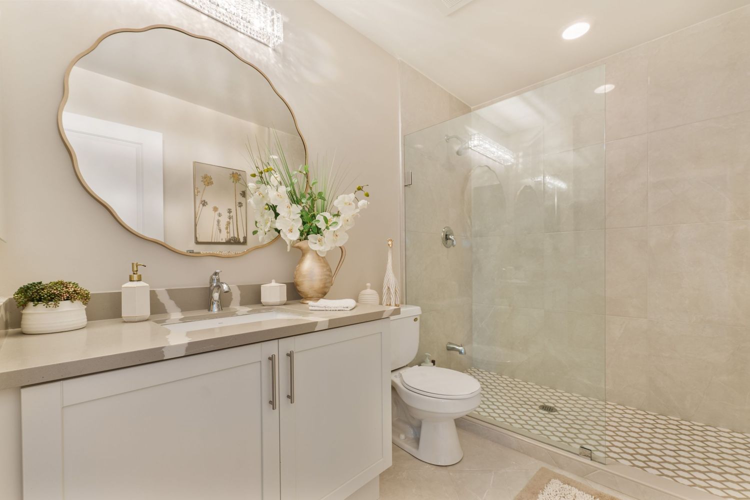 Elegant bathroom with white vanity, round gold mirror, glass shower, and fresh floral arrangement in neutral tones.