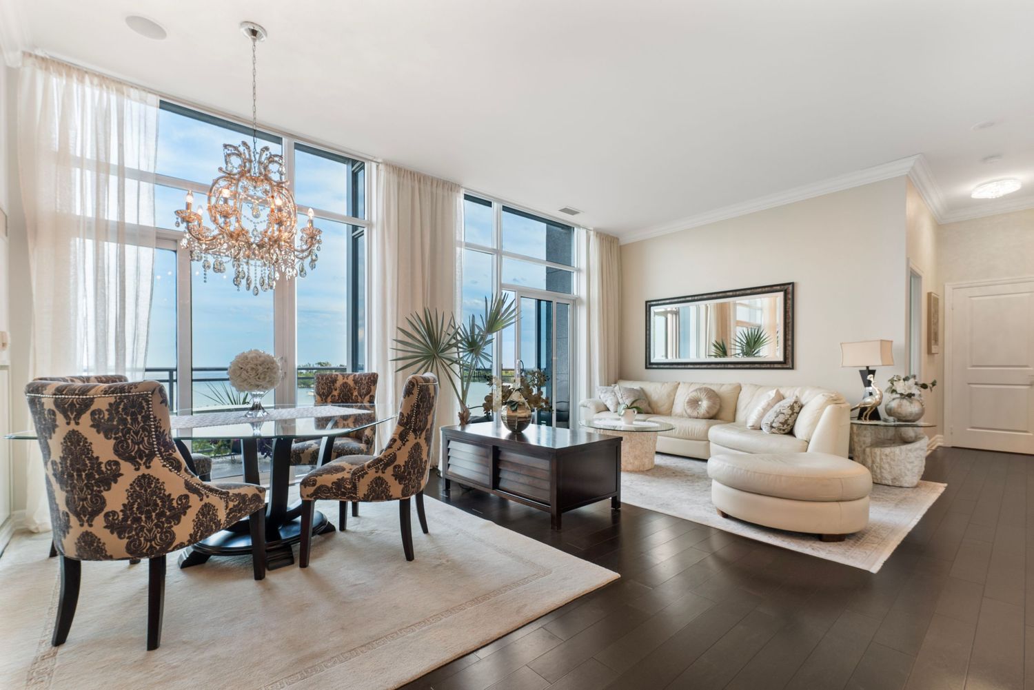 Elegant dining and living room with chandelier, floor-to-ceiling windows, dark hardwood floors, and luxurious neutral decor.