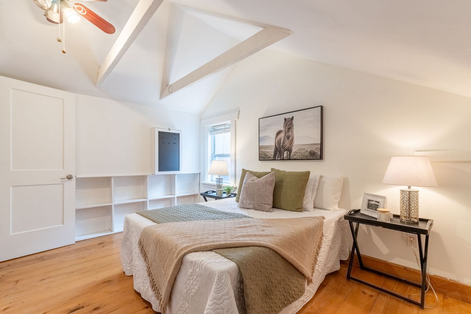 Bright bedroom with vaulted ceiling, built-in shelving, hardwood floors, and horse artwork above the bed.