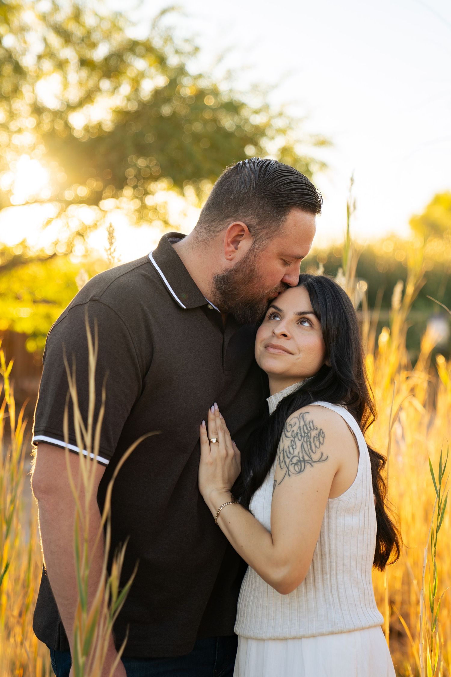 Couple shares tender moment at golden sunset in tall grass field, man kissing tattooed womans temple. Las Vegas portrait photographer