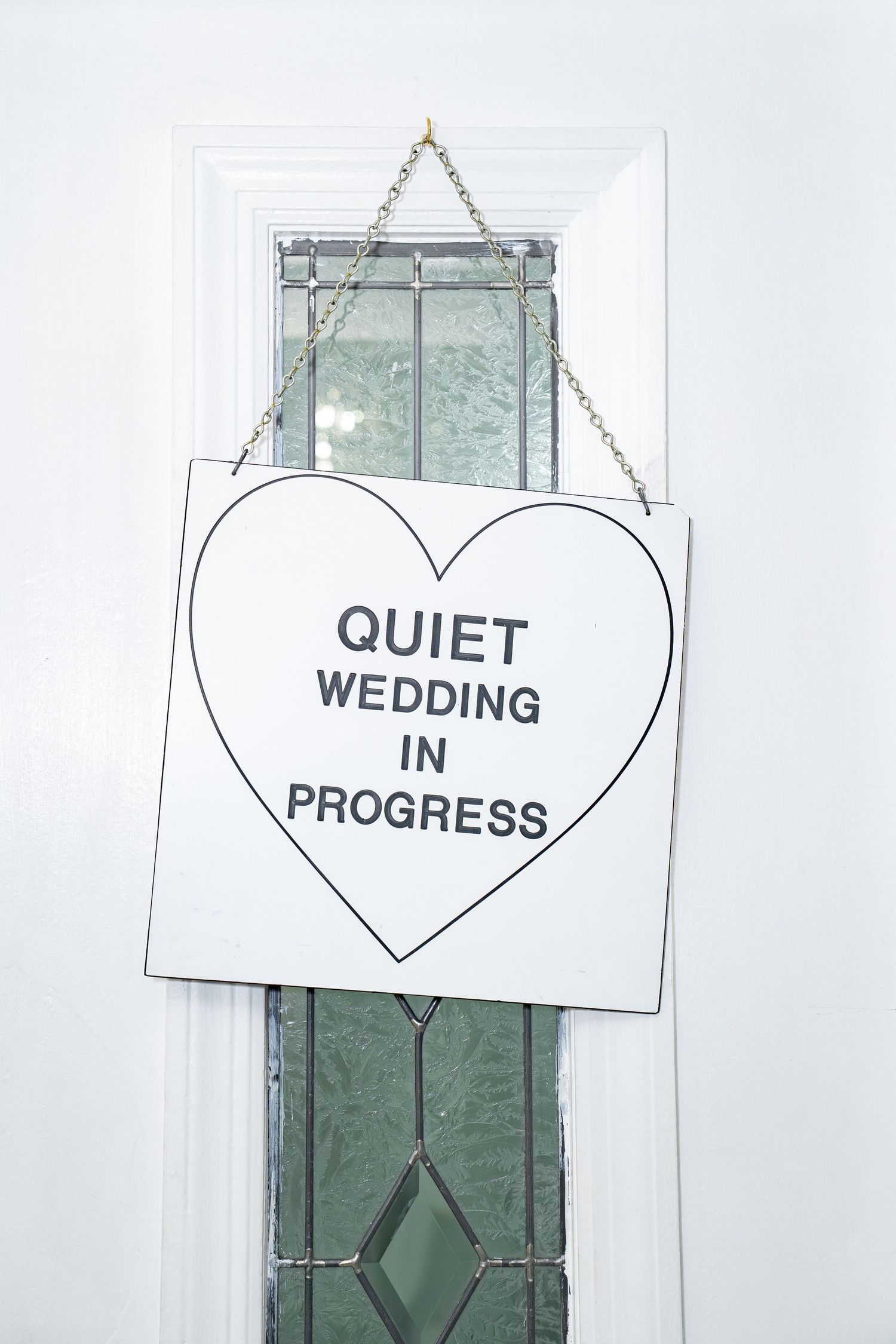 Heart-shaped sign reading Quiet Wedding In Progress hanging on white chapel door with stained glass window. Las Vegas elopement photographer