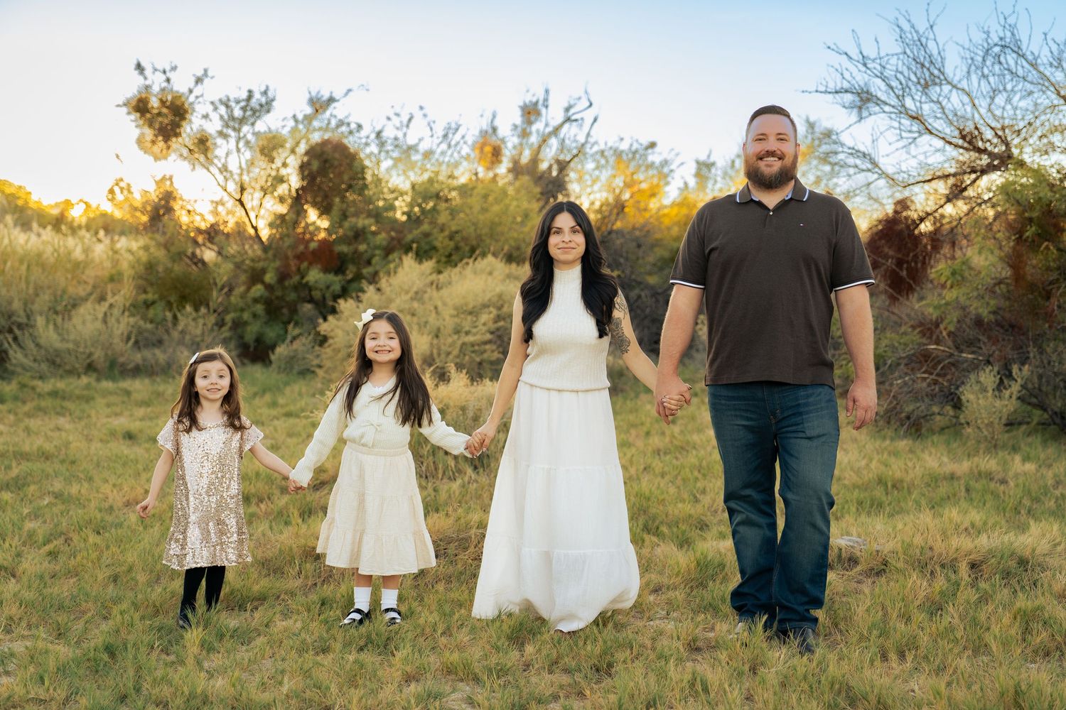 Family of four holds hands walking through scenic desert meadow at sunset in coordinated white outfits. Las Vegas portrait photographer