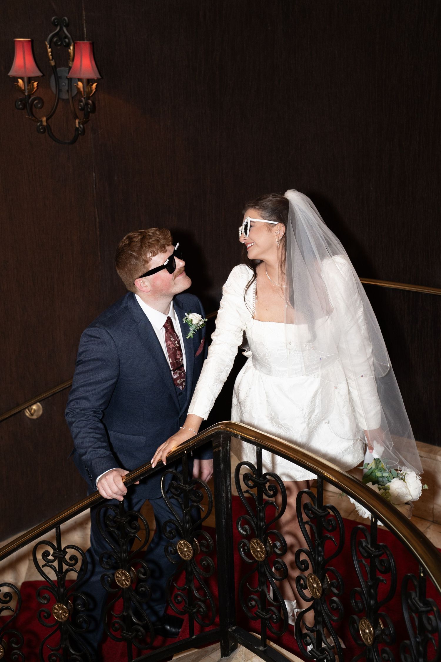 Newlyweds in sunglasses pose on ornate staircase, bride in short white dress with veil, groom in navy suit. Las Vegas elopement photographer