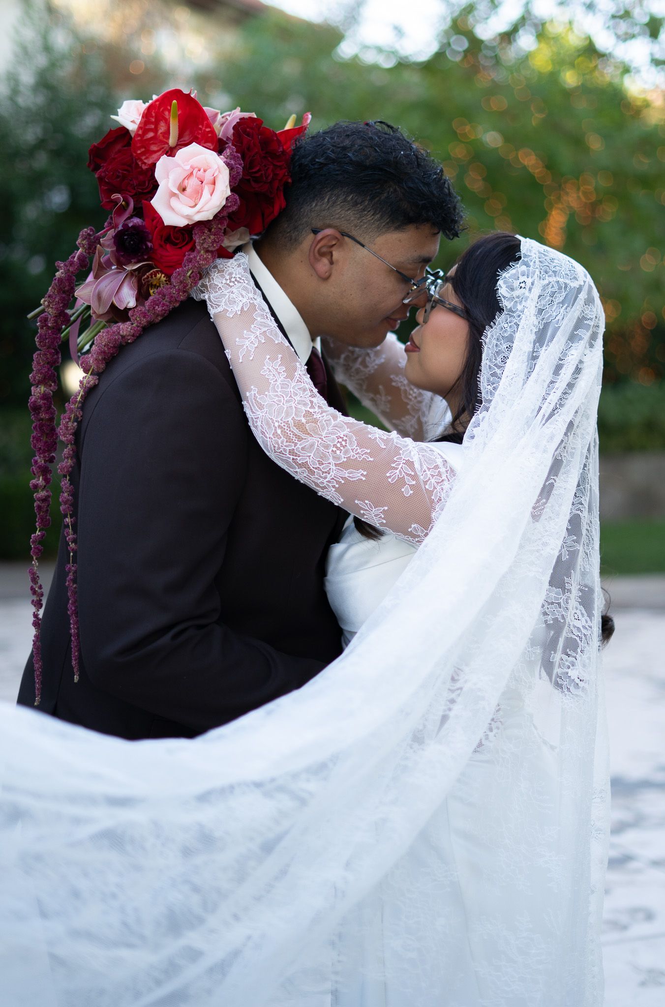 Couple shares romantic kiss during outdoor wedding, bride in lace veil adorned with red and pink flowers. Las Vegas wedding photographer