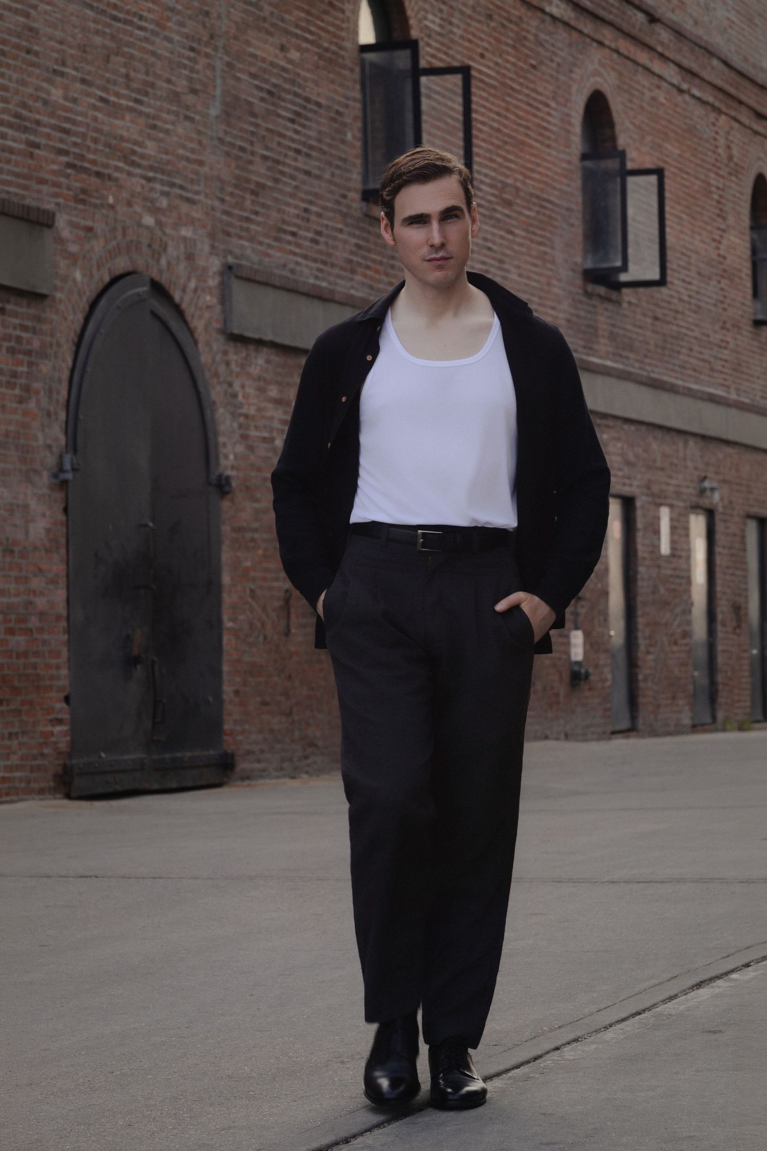 Full-length portrait of Dylan walking toward the camera across an industrial courtyard lined with arched warehouse doors in Red Hook.