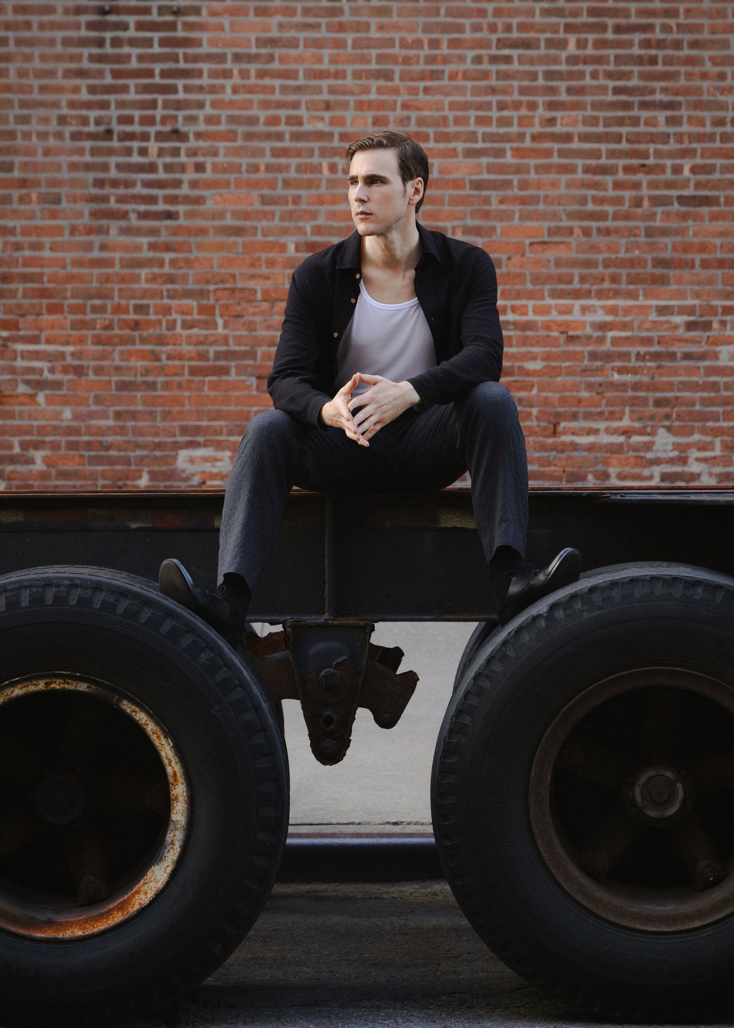 Dylan sits between two large trailer tires and looks off frame in a symmetrical industrial portrait against a brick wall.