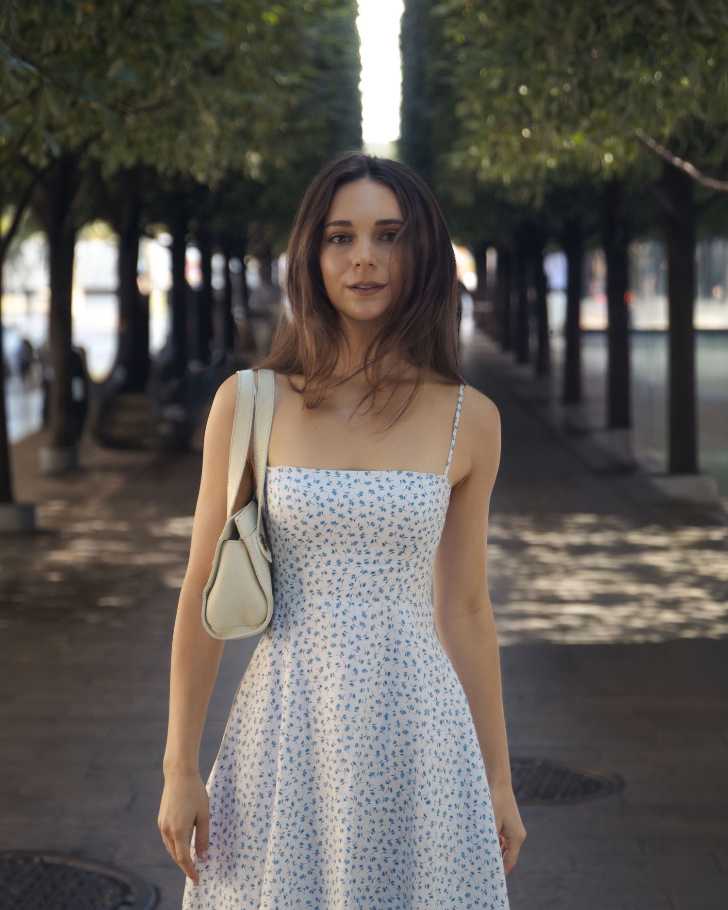 Portrait of Anastasia in a floral dress beneath two rows of trees, with a cream handbag over her shoulder.