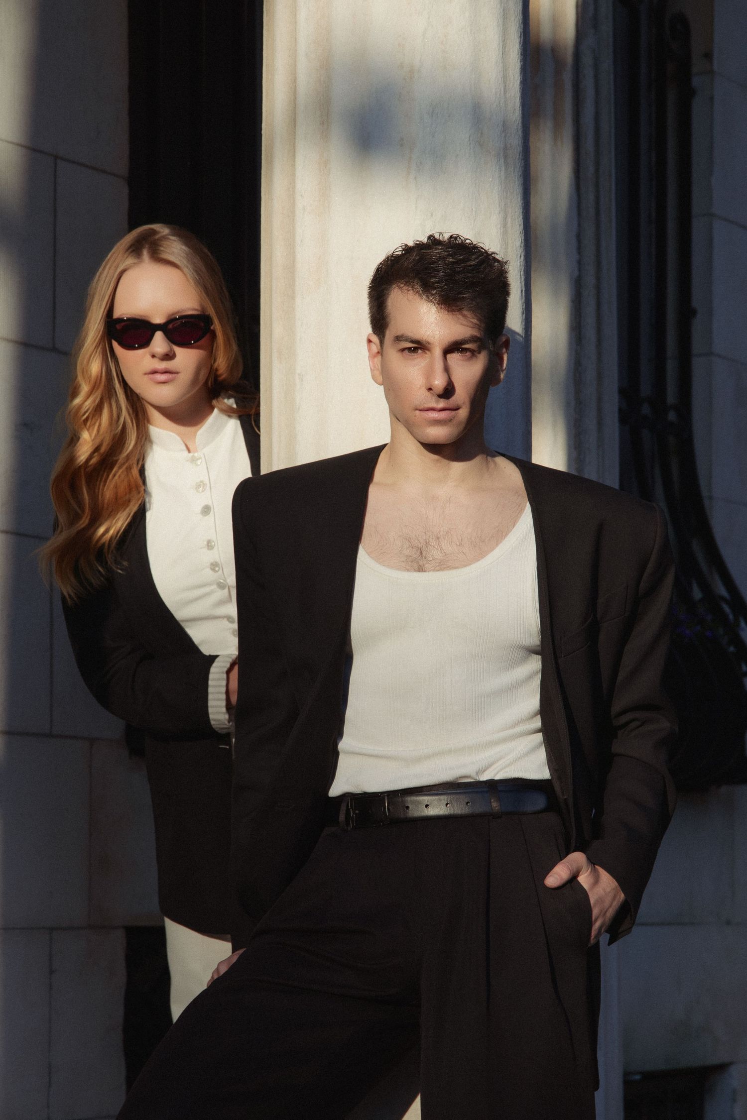 Vin poses in the foreground while Regina stands behind him in sunglasses, both framed by sharp sunlight and deep architectural shadows.