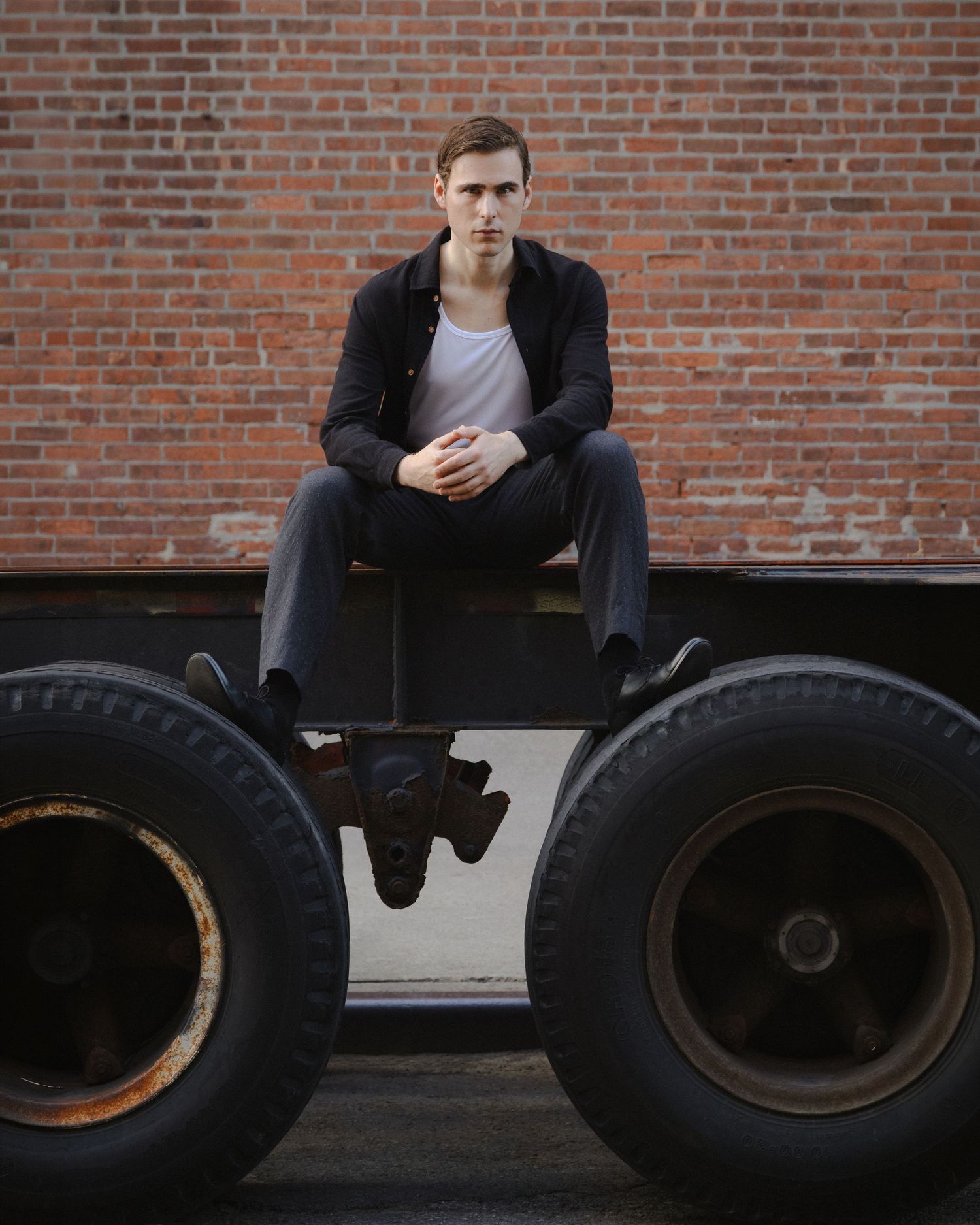 Dylan sits on a metal trailer frame between two oversized truck tires, centered against a red brick wall in Red Hook, Brooklyn.