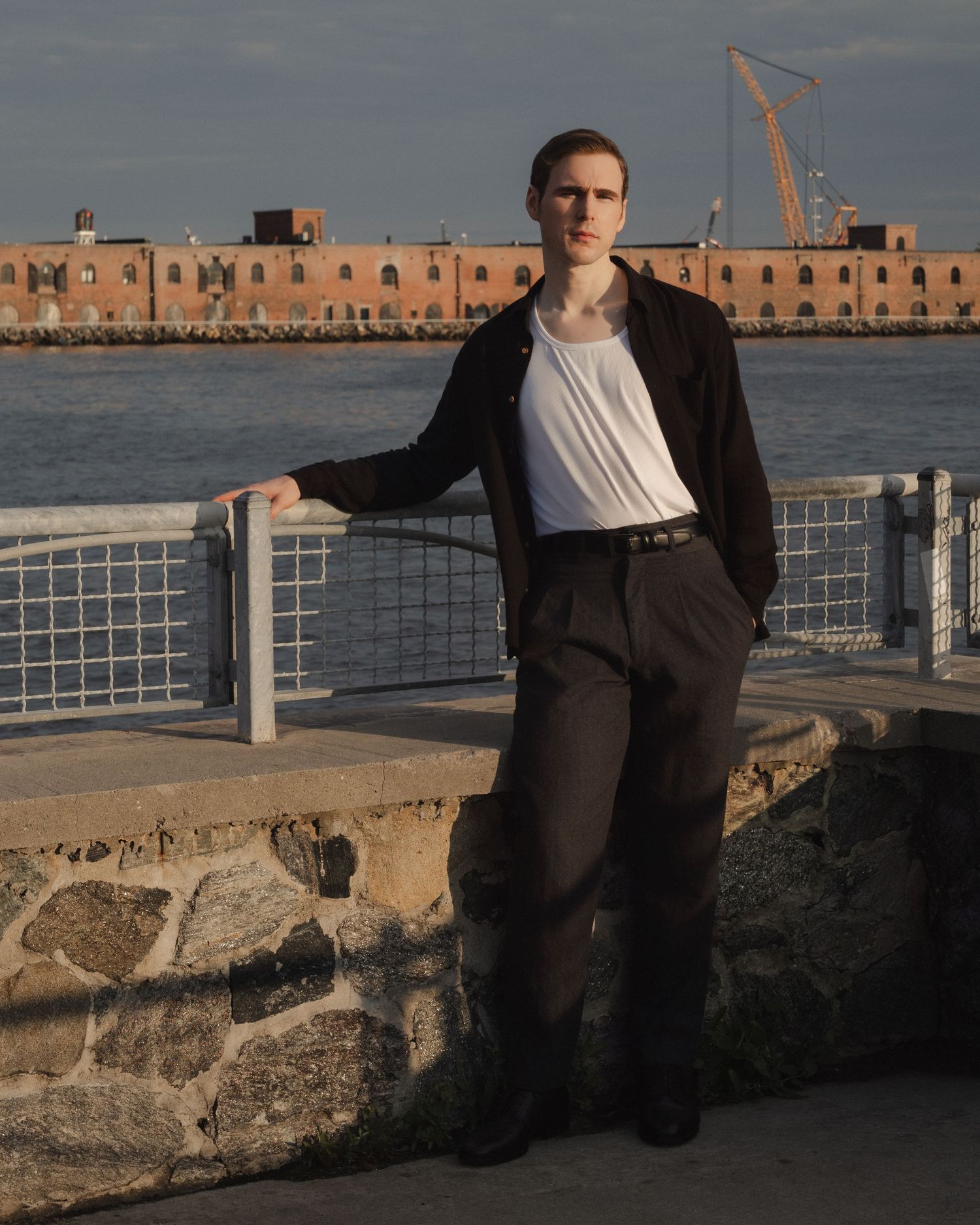 Dylan leans against a waterfront railing in warm sunlight, with a brick warehouse and construction crane across the water.