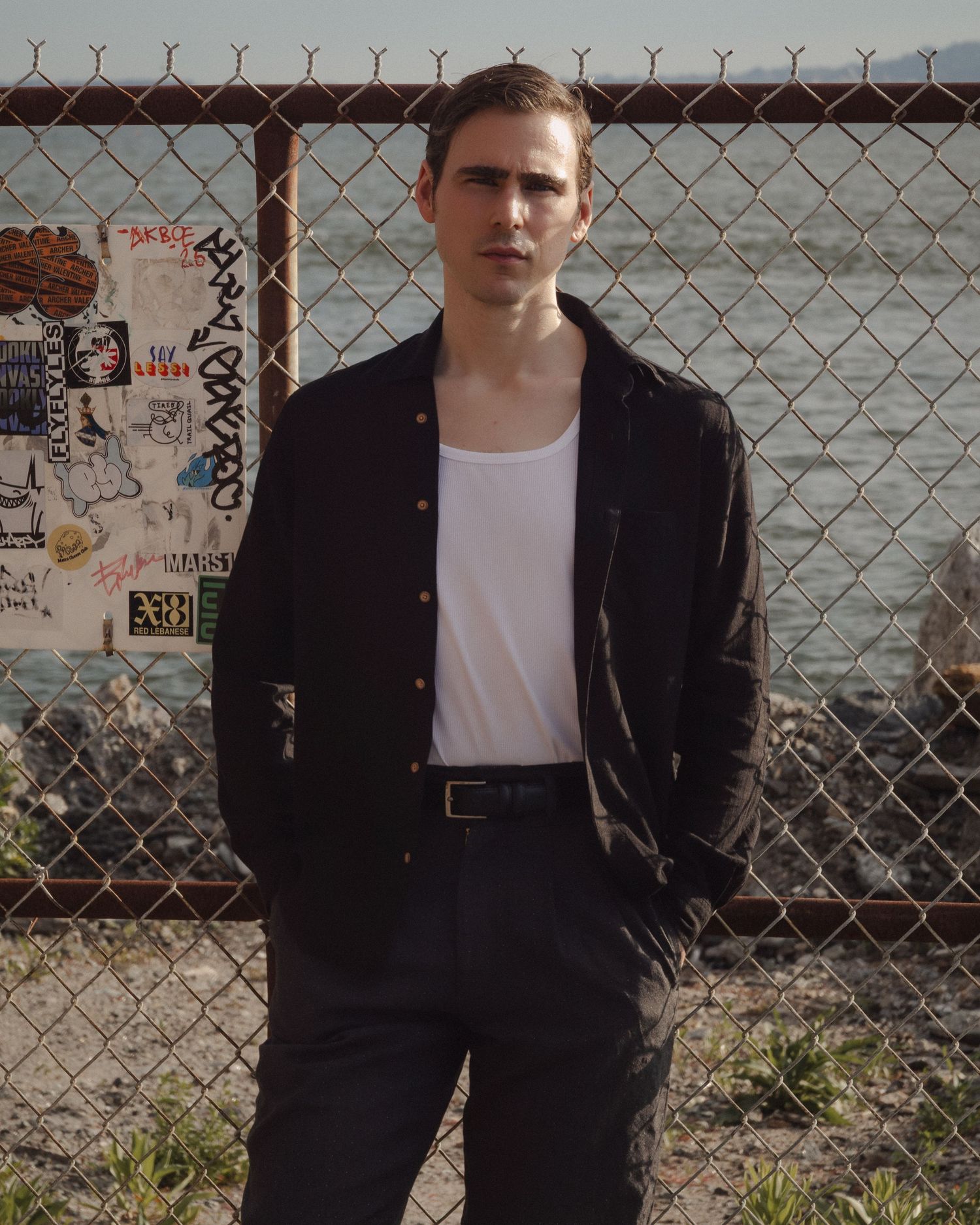 Dylan stands before a chain-link fence and sticker-covered panel, with the rocky waterfront visible beyond.