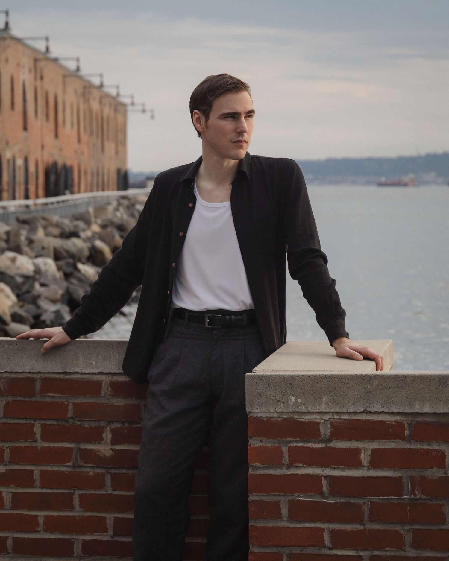 Dylan rests both hands on a brick waterfront wall, framed by an industrial warehouse, rocky shoreline, and open water.