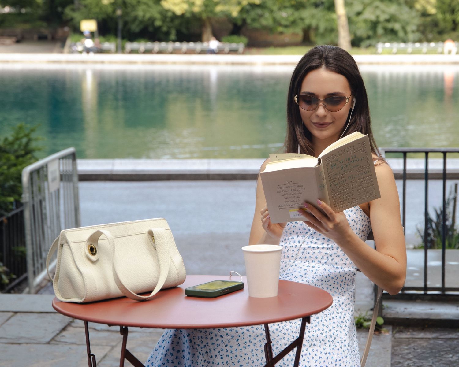Woman in sunglasses reading a book at a café table overlooking the water in Central Park.