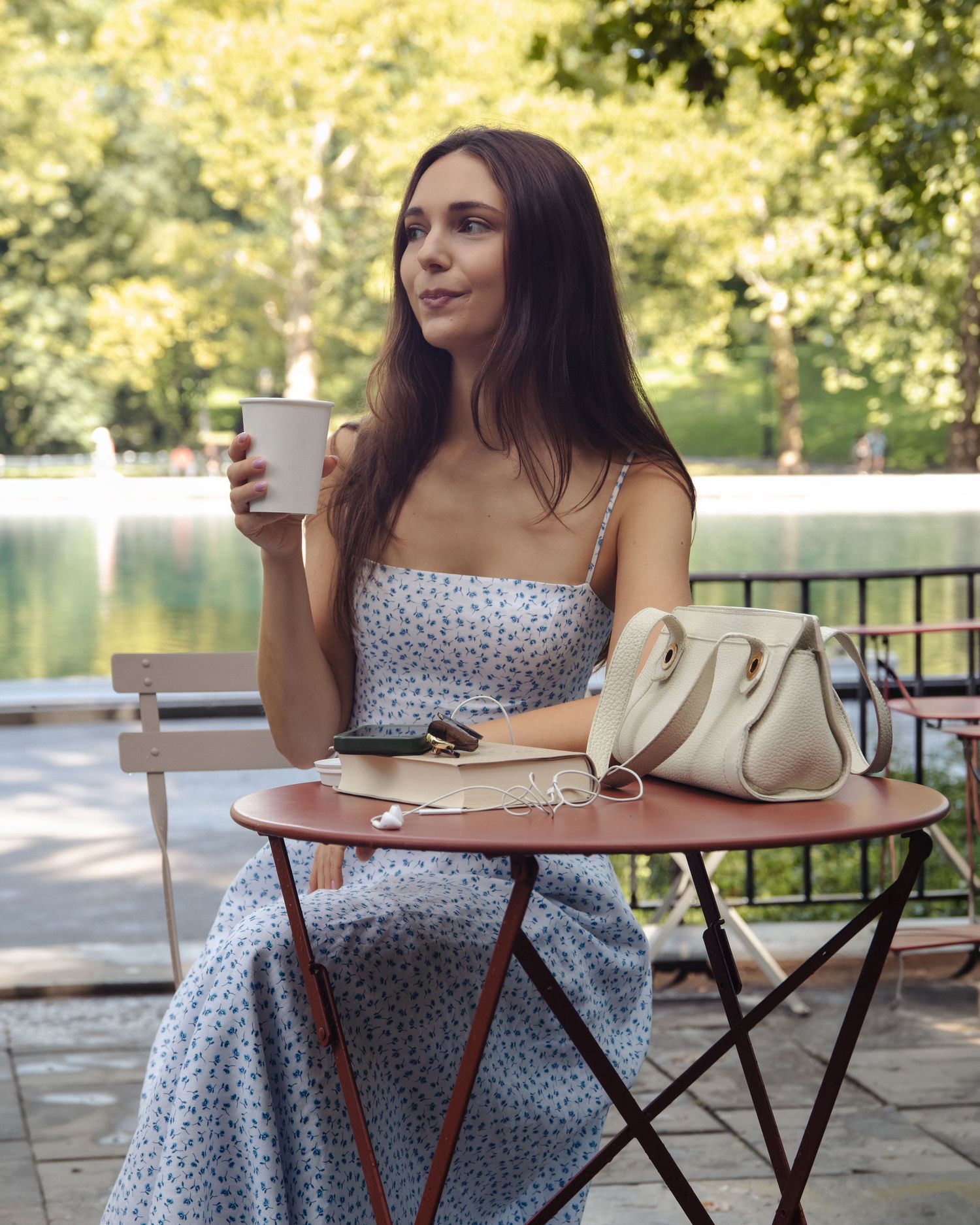 Woman holding a coffee cup at an outdoor café beside the water, with a book and cream handbag on the table.