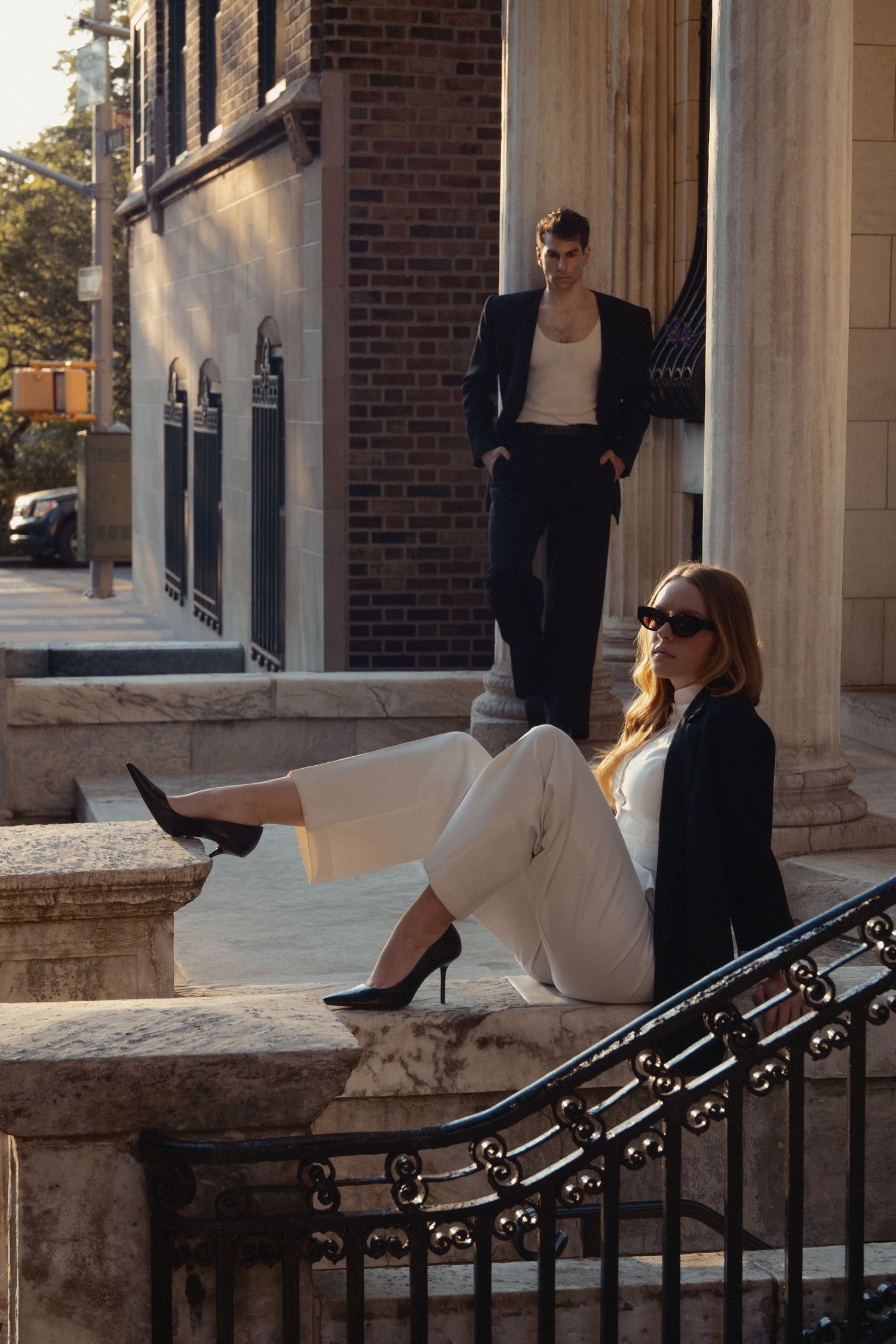 Regina sits in sunglasses on a stone ledge while Vin stands behind her among columns and wrought-iron railings on the Upper West Side.