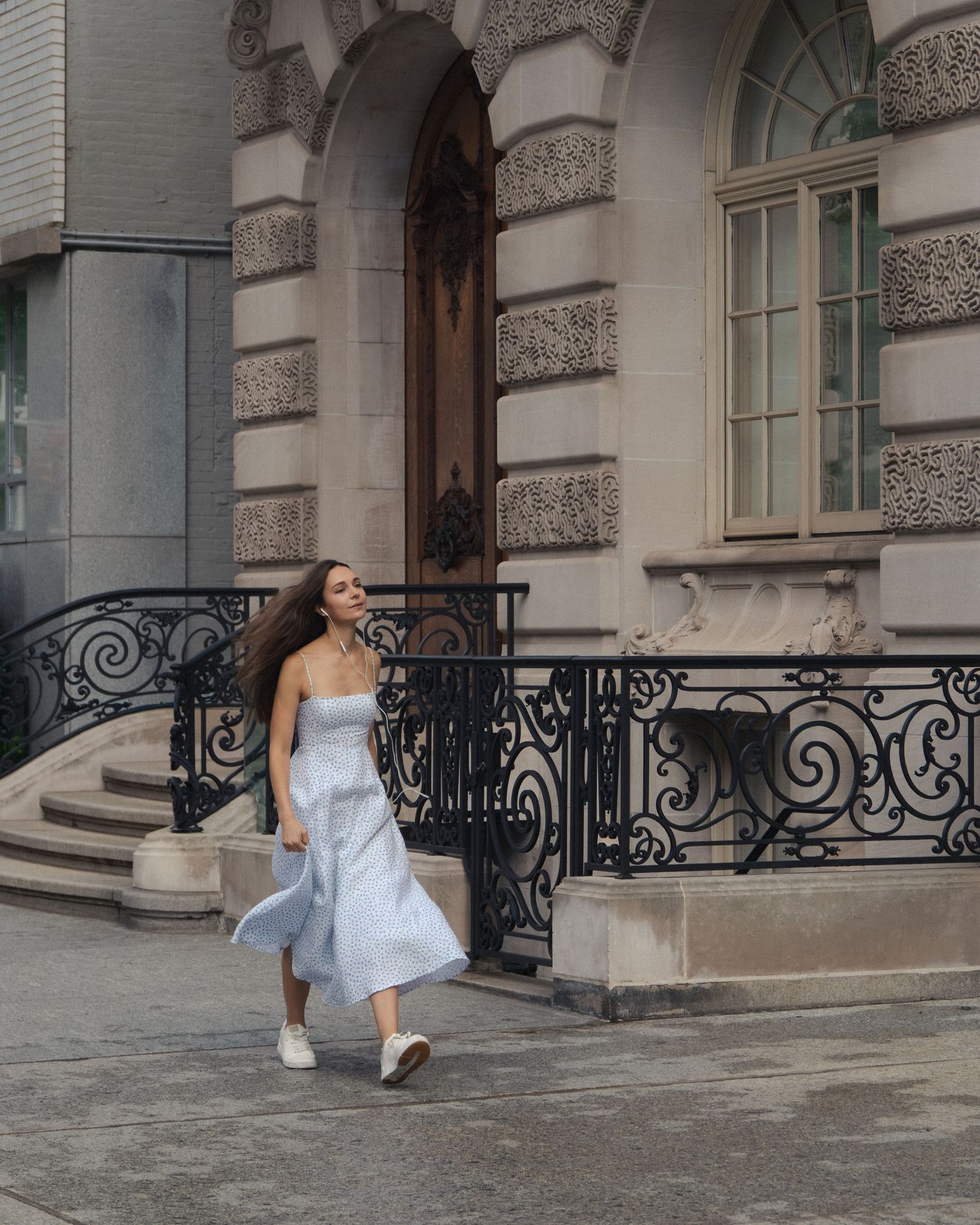 Woman walking past an ornate stone townhouse and wrought-iron railings, her hair and floral skirt flowing behind her.