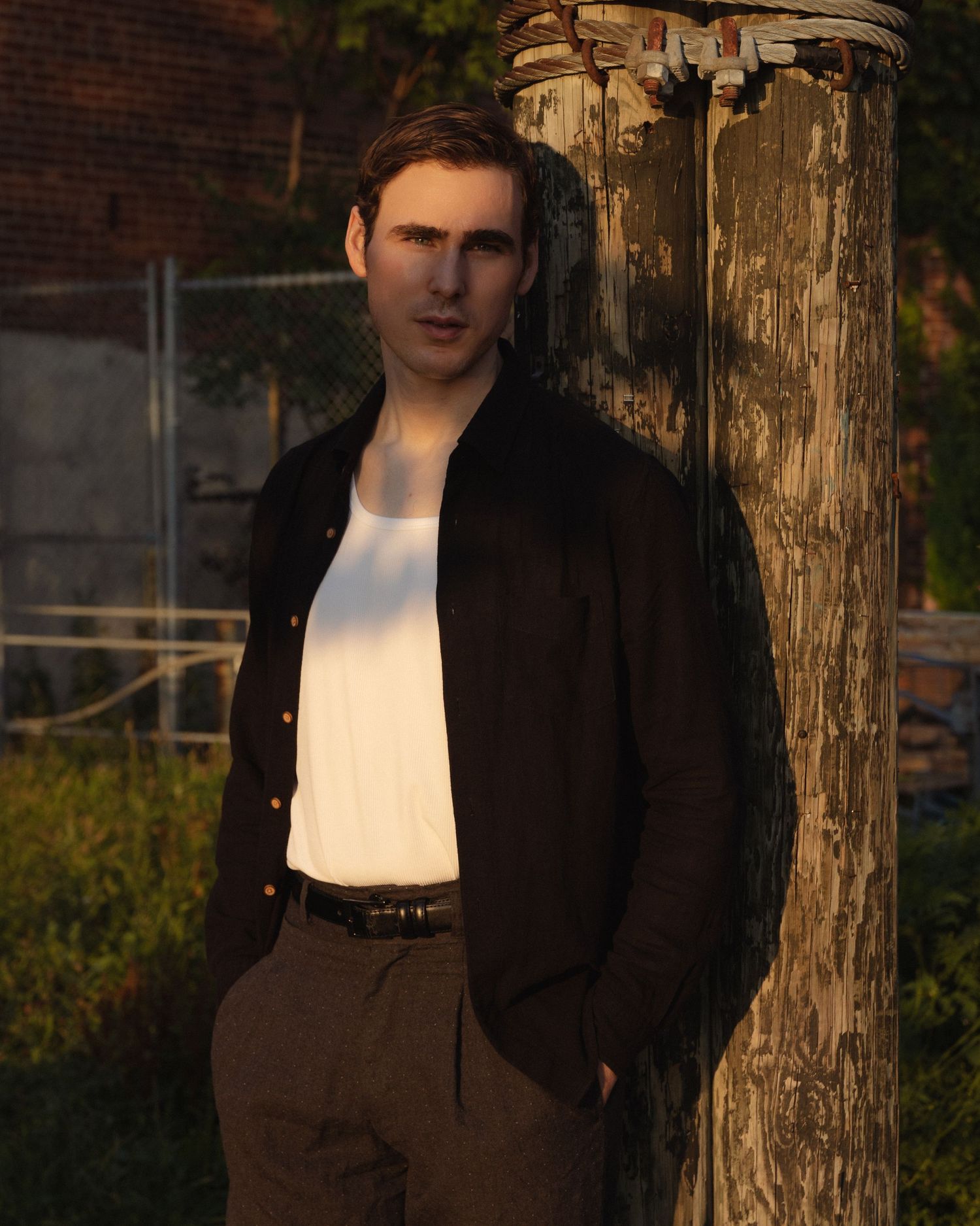 Dylan leans against a weathered wooden post wrapped with rope, his face crossed by warm late-day sunlight.