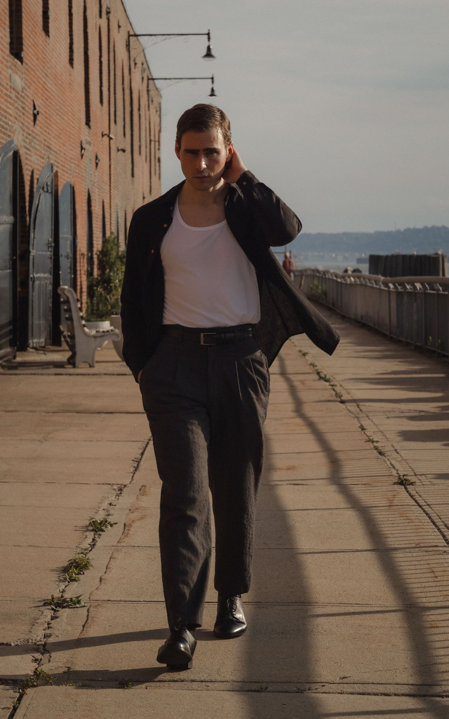 Dylan walks along a sunlit Red Hook waterfront path beside a long brick warehouse, with the harbor visible behind him.