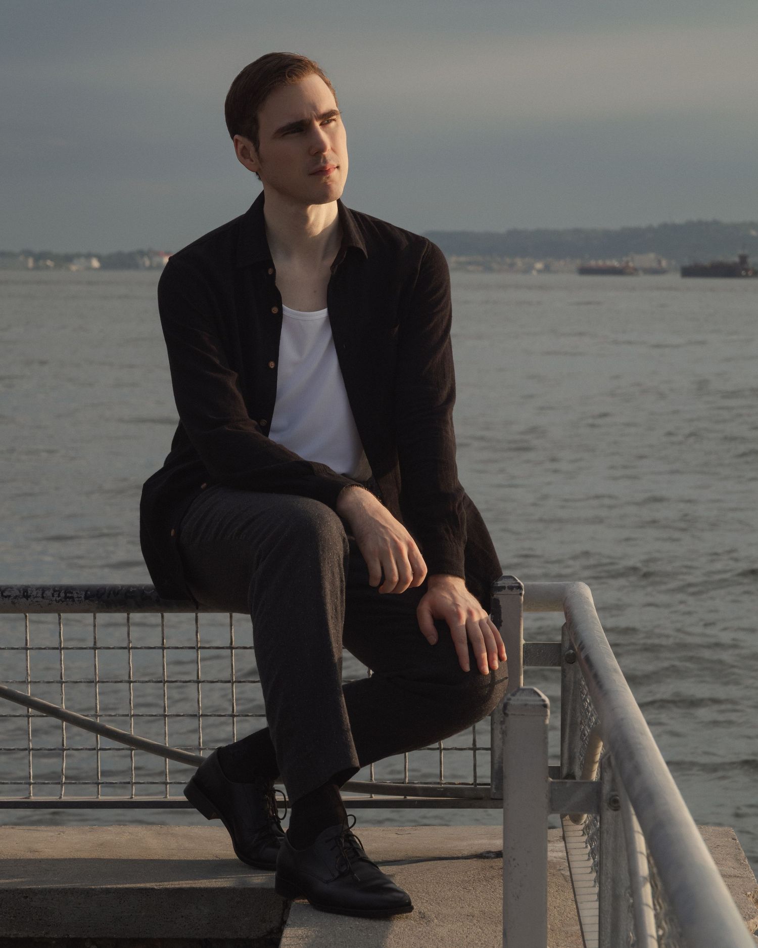 Dylan sits on a metal harbor railing above the water in a quiet Red Hook waterfront portrait.