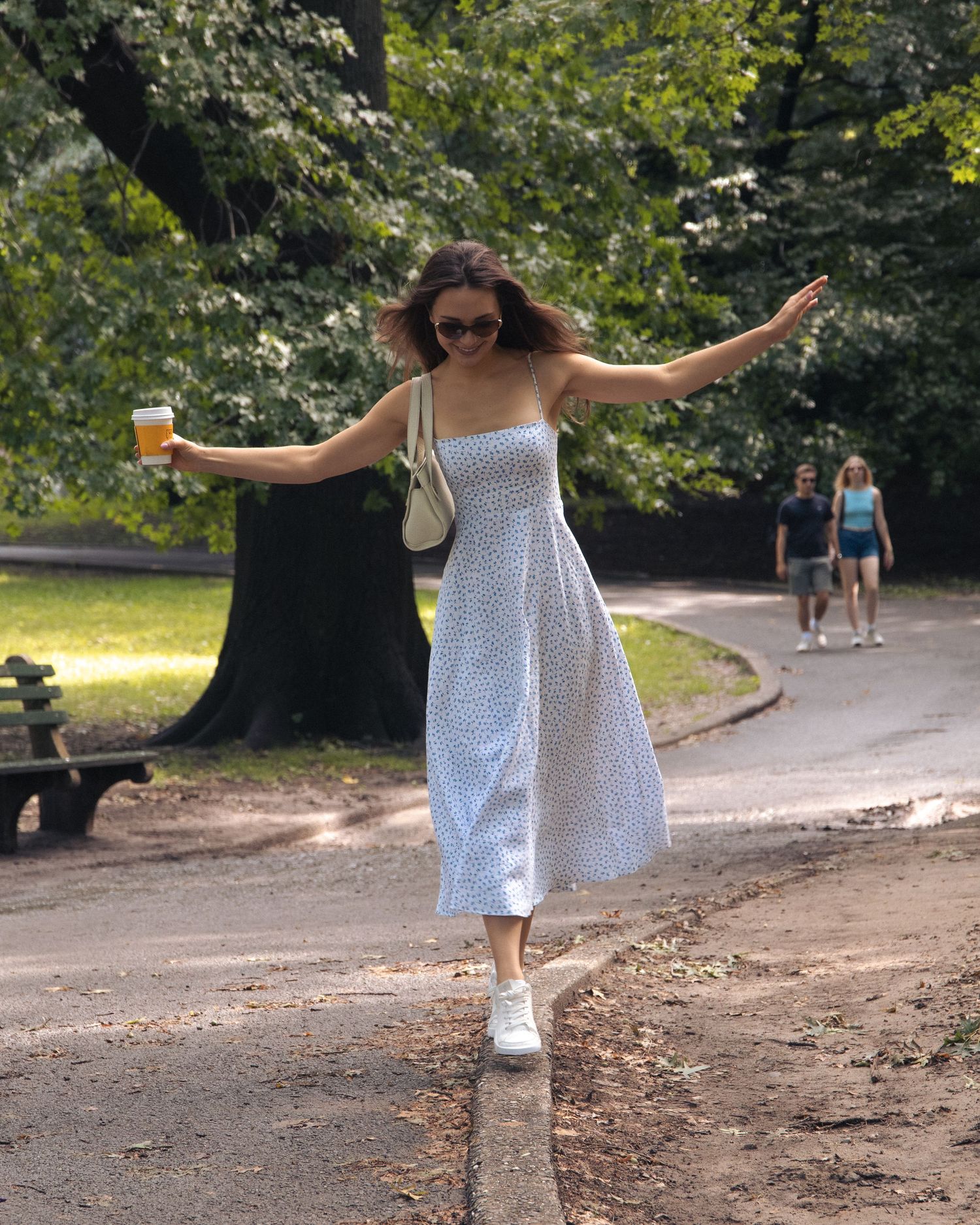 Woman balancing on a curb along a Central Park path, arms outstretched and a takeaway coffee in one hand.